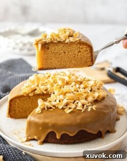 A hand holding a slice of moist caramel mud cake above the remaining cake, highlighting its rich texture and perfect portioning, ready to be served.