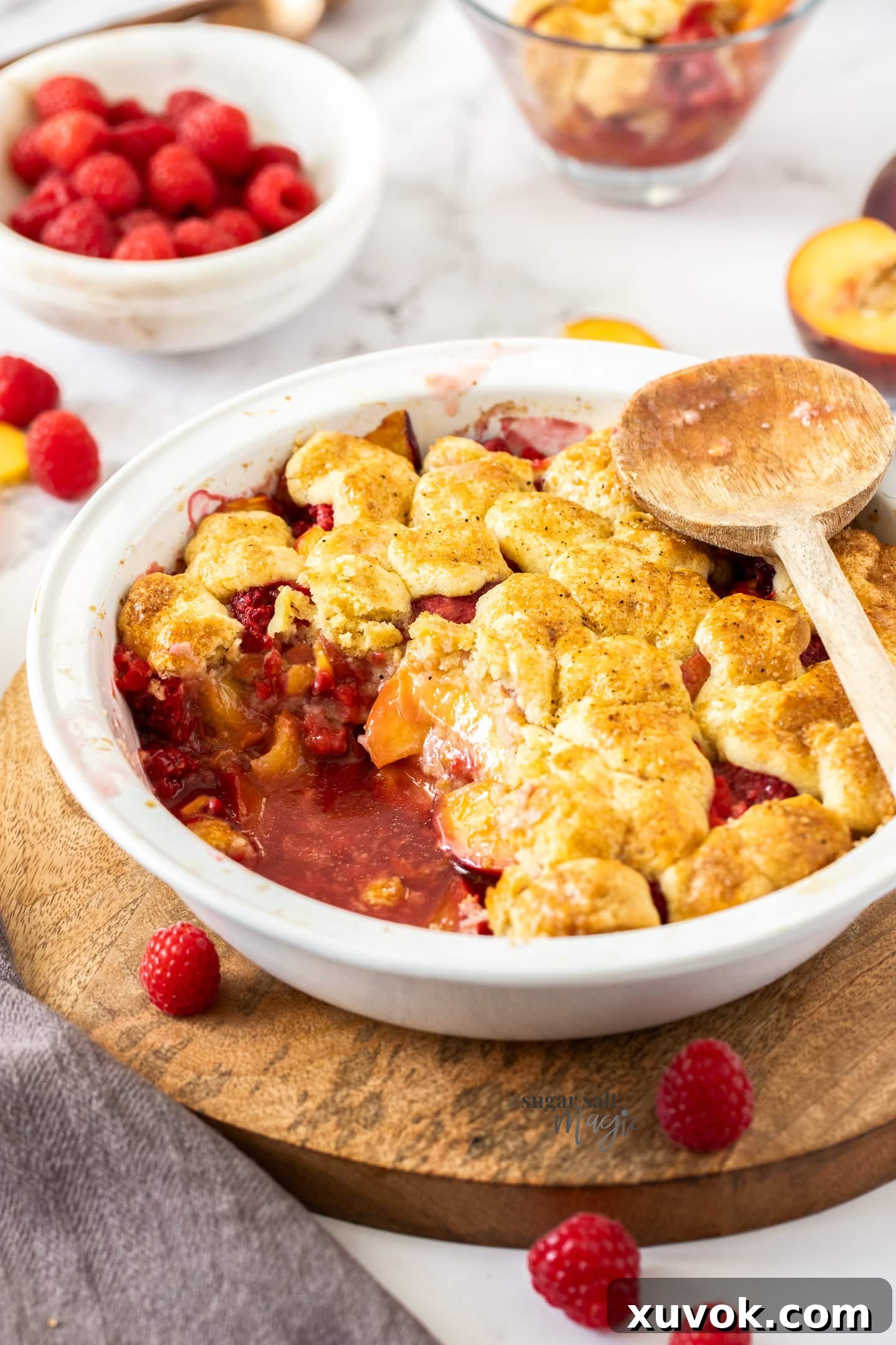 A beautifully baked raspberry peach cobbler in a white pie dish on a wooden platter, ready to be served.
