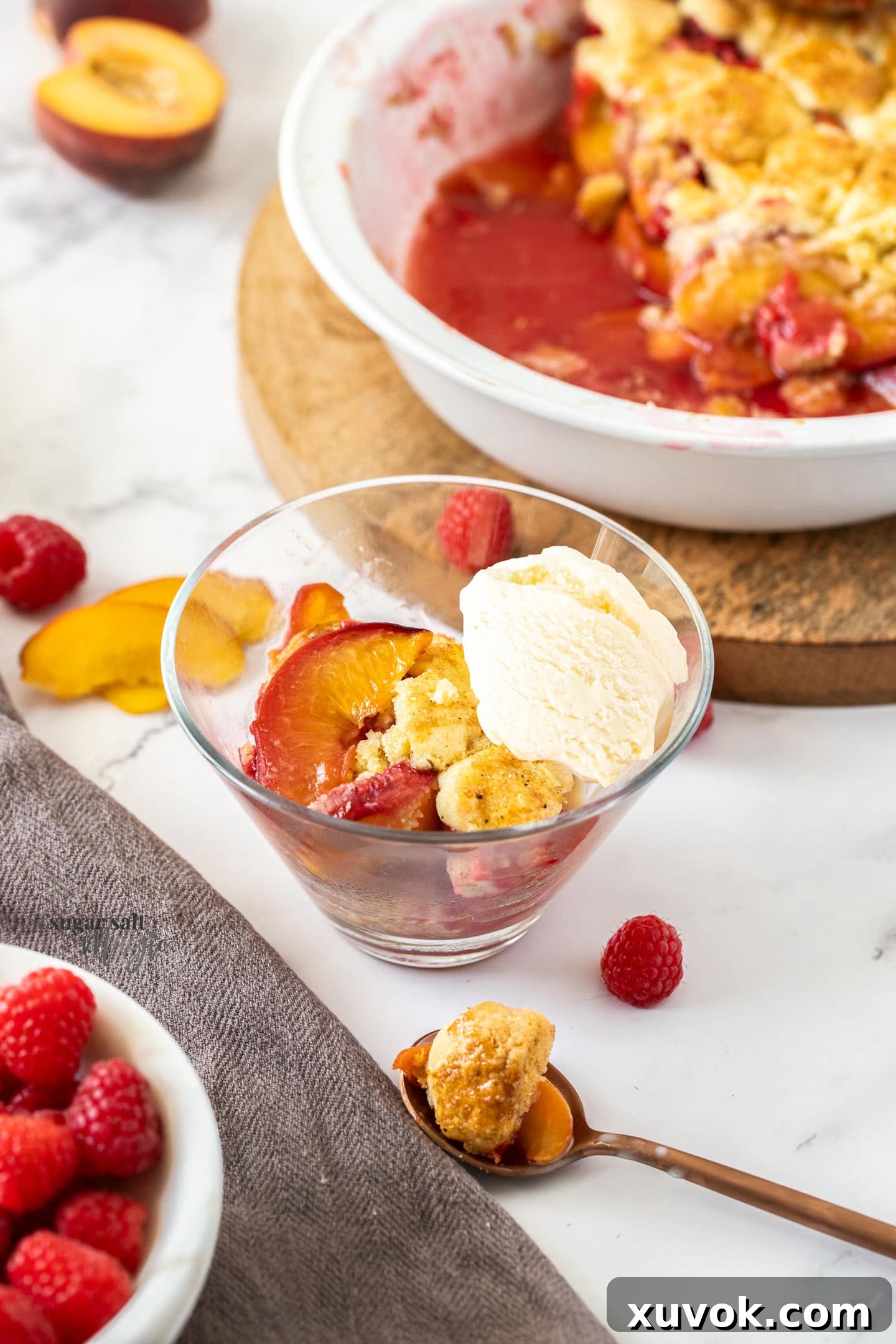 A serving of warm raspberry peach cobbler in a clear glass bowl, topped with a scoop of melting vanilla ice cream, on a rustic wooden surface.
