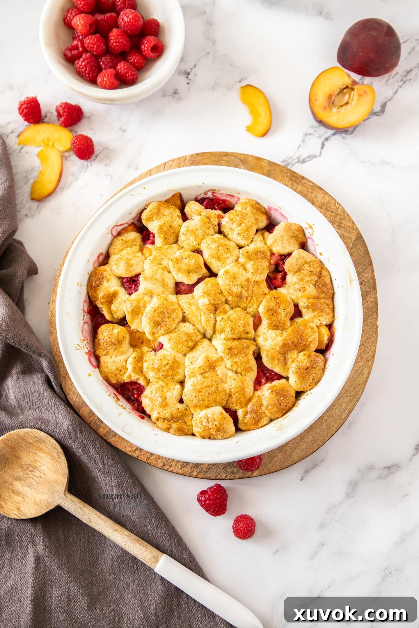 A top-down view of a warm raspberry peach cobbler, showcasing the golden biscuit topping with hints of bright red raspberries peeking through.
