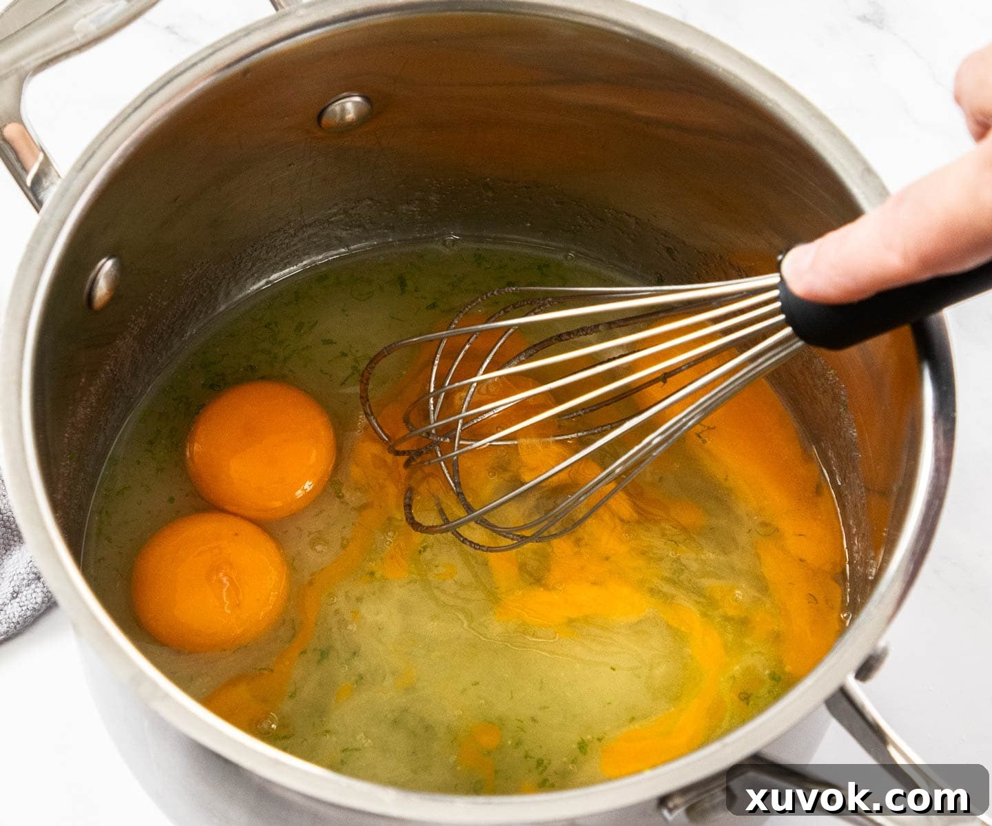 A whisk blending eggs, sugar, and lime juice in a saucepan, creating the initial base for the curd.