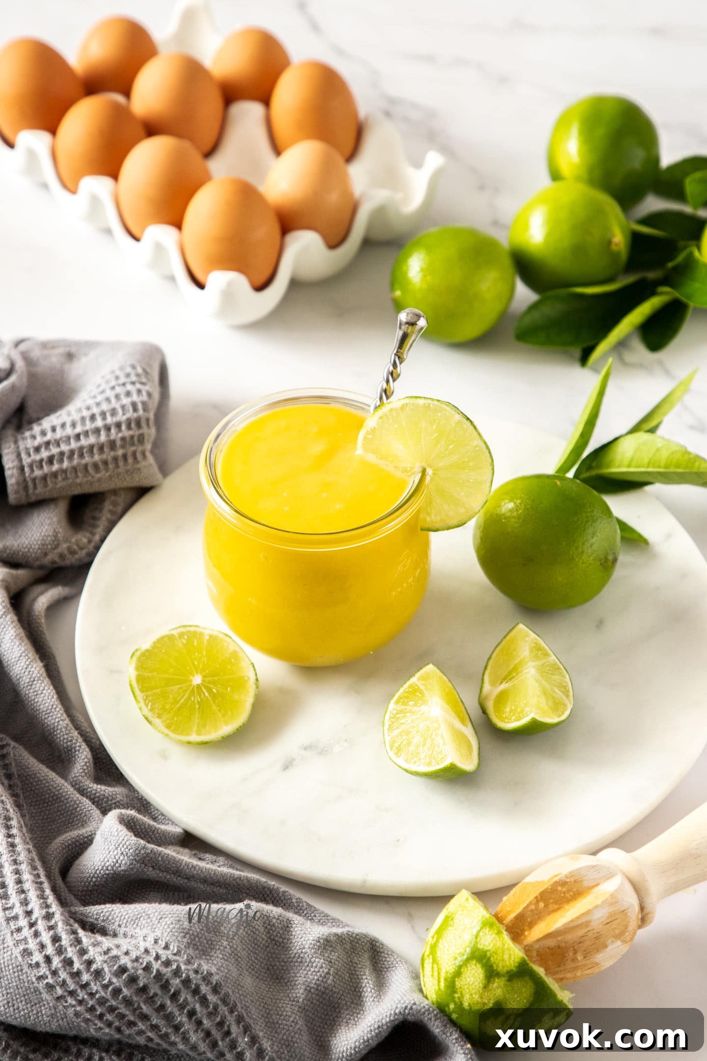 A beautifully presented glass jar of lime curd on a white platter, accented by fresh limes and whole eggs.