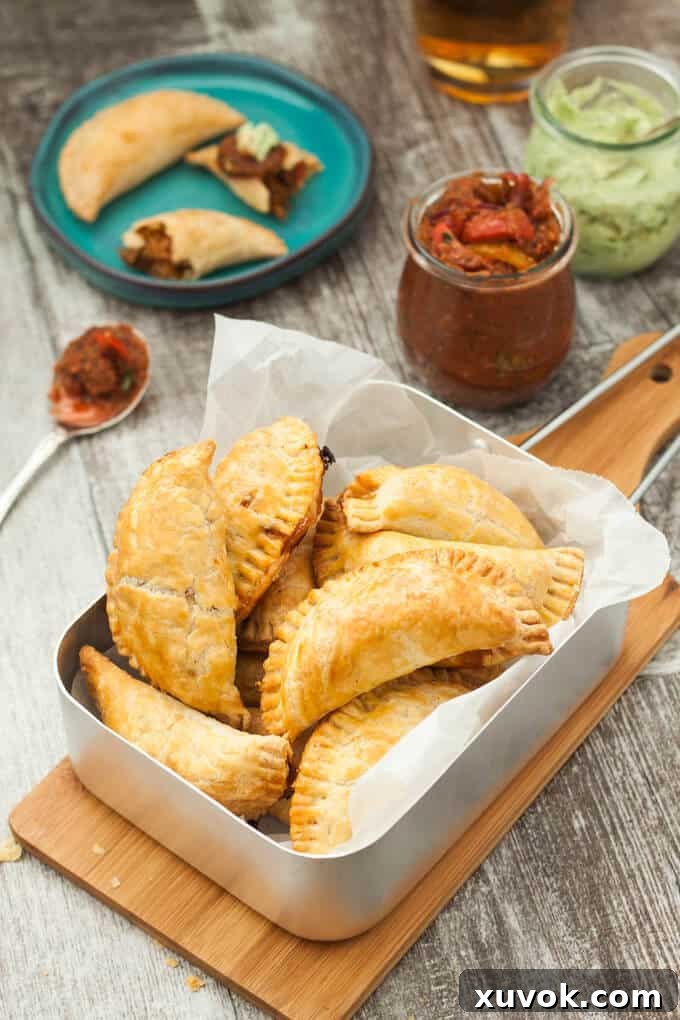 Golden brown pulled pork empanadas arranged neatly in a steel baking tray, with various dipping sauces blurred in the background, suggesting a perfect appetizer or snack spread.