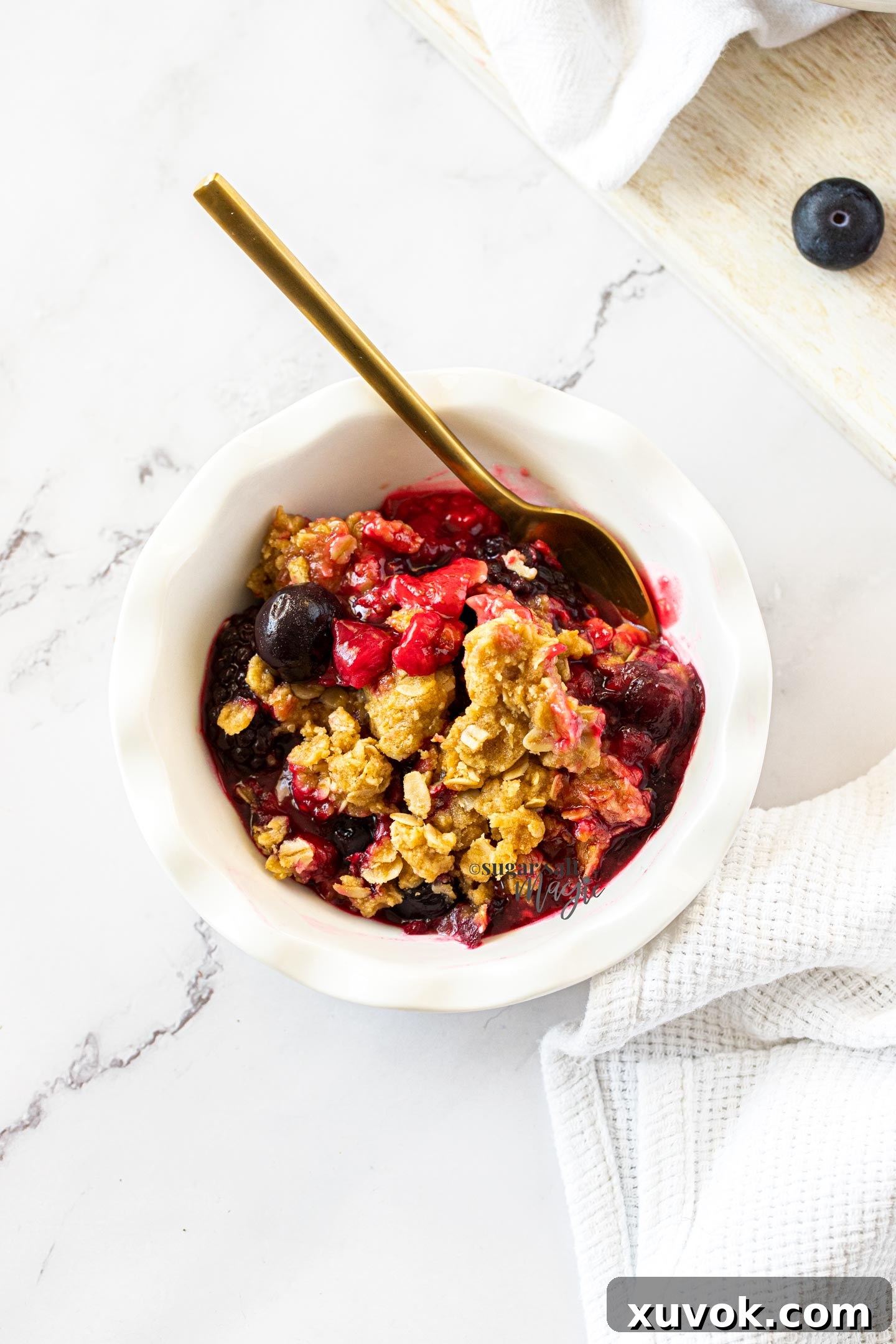 A miniature white pie dish filled with baked crumble and berries, with a single golden spoon resting in it.