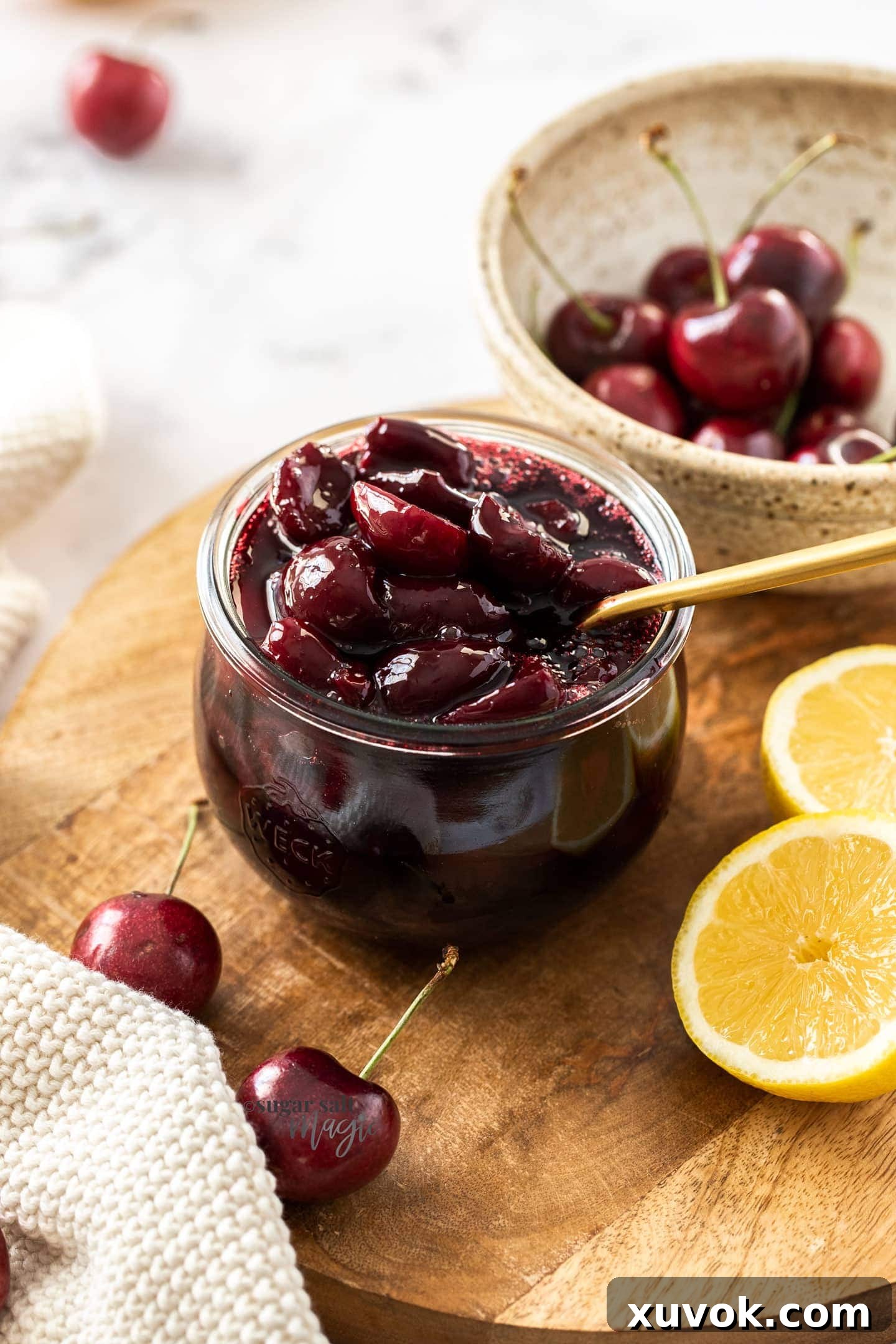 A glass jar filled with cherry compote with a gold spoon in it. Sitting on a wooden board