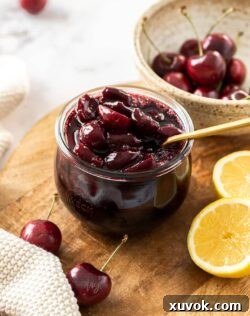 A glass jar filled with cherry compote with a gold spoon in it. Sitting on a wooden board.