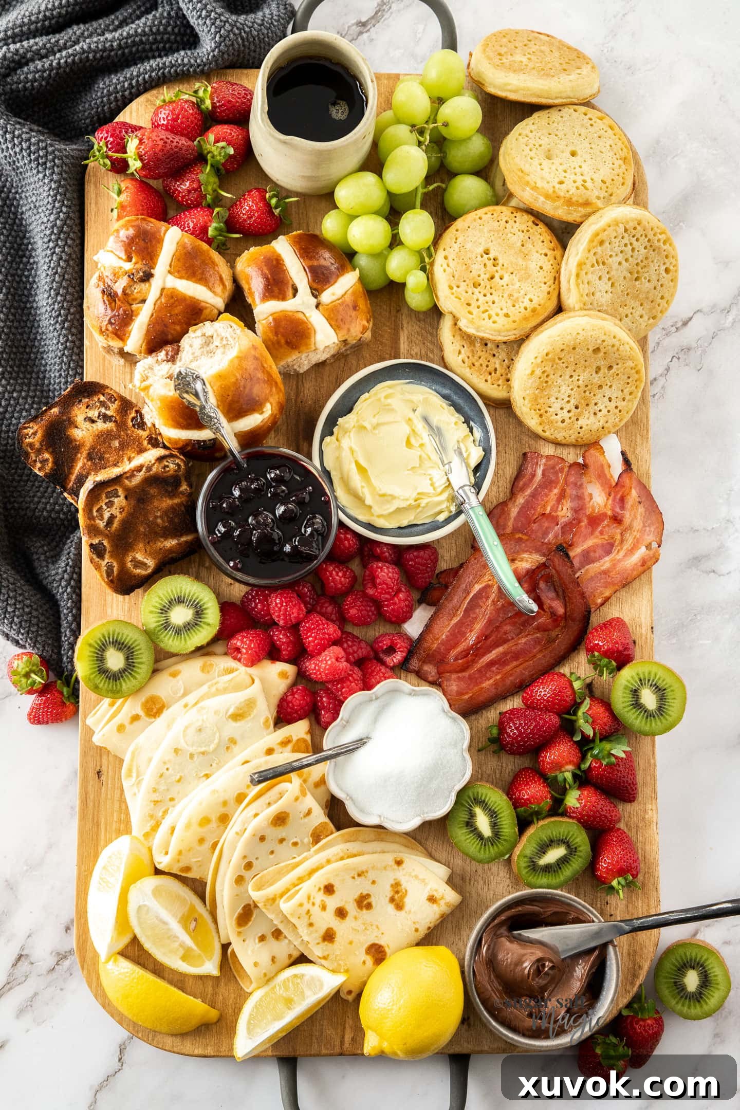 A wooden board filled with a variety of breakfast foods, including pancakes, crumpets, fruits, and condiments.