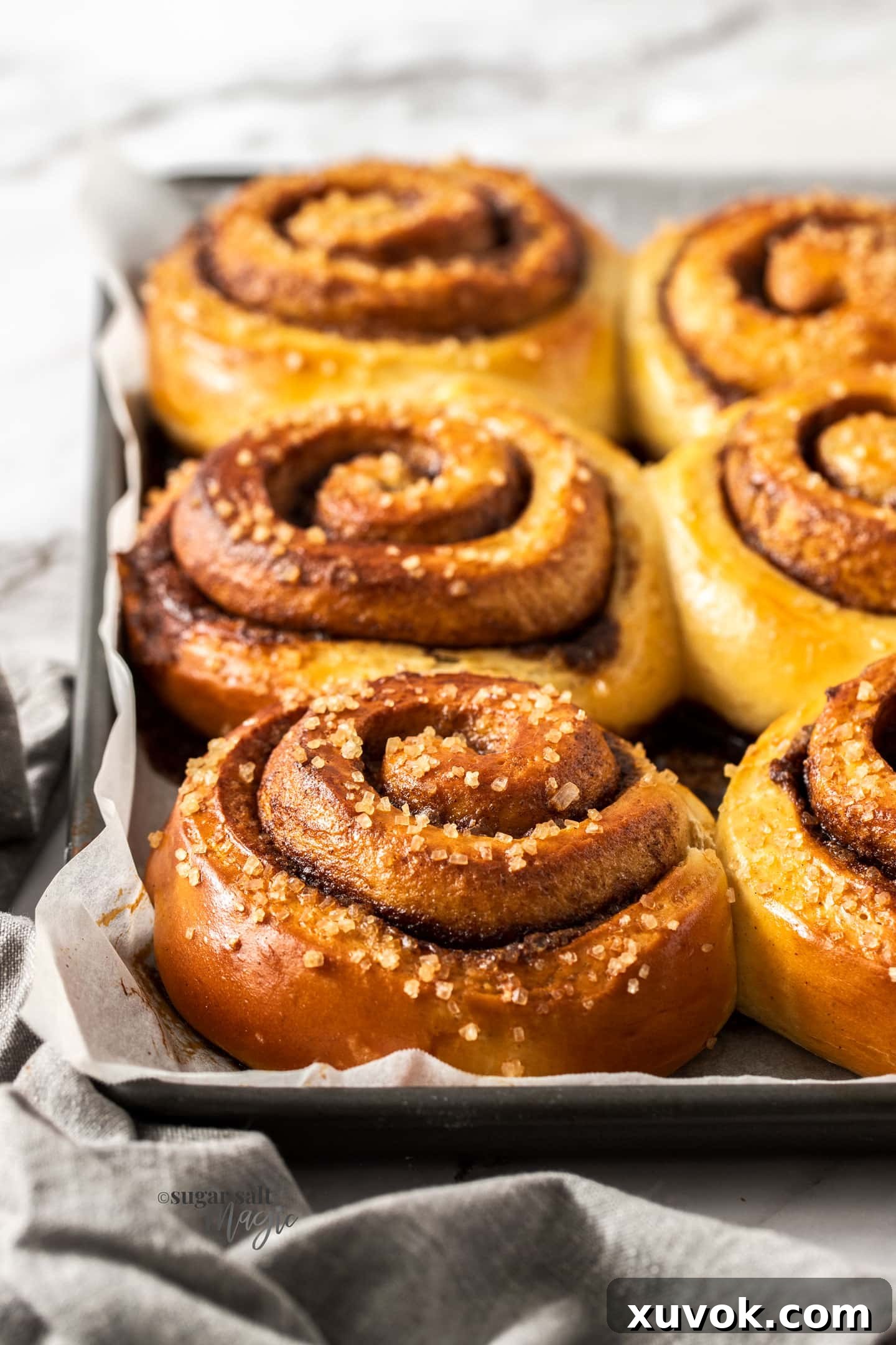 A row of 3 jumbo Swedish cinnamon buns on a baking tray, showcasing their golden-brown tops and pearl sugar sprinkles.