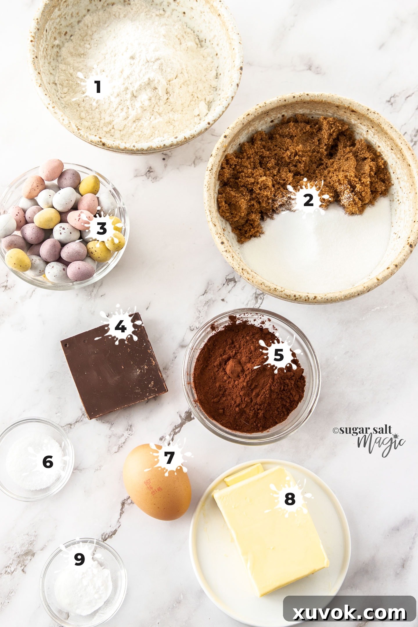 Various baking ingredients neatly arranged on a white surface, including flour, sugars, butter, cocoa, eggs, and Cadbury mini eggs, ready for making chocolate Easter egg cookies.