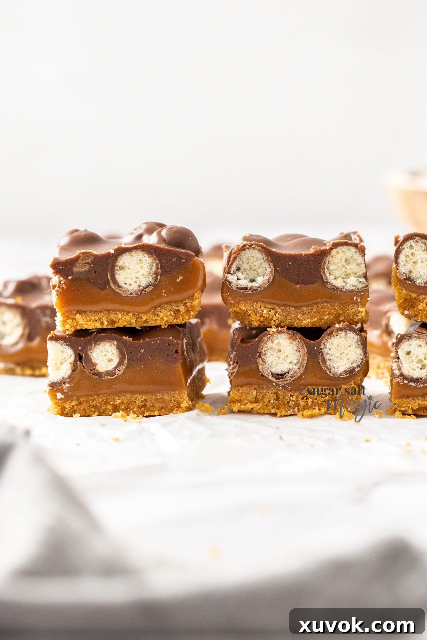 Two stacks of beautifully cut Caramel Malteser Slices on baking paper, showcasing their distinct layers of biscuit base, caramel, Maltesers, and chocolate ganache.
