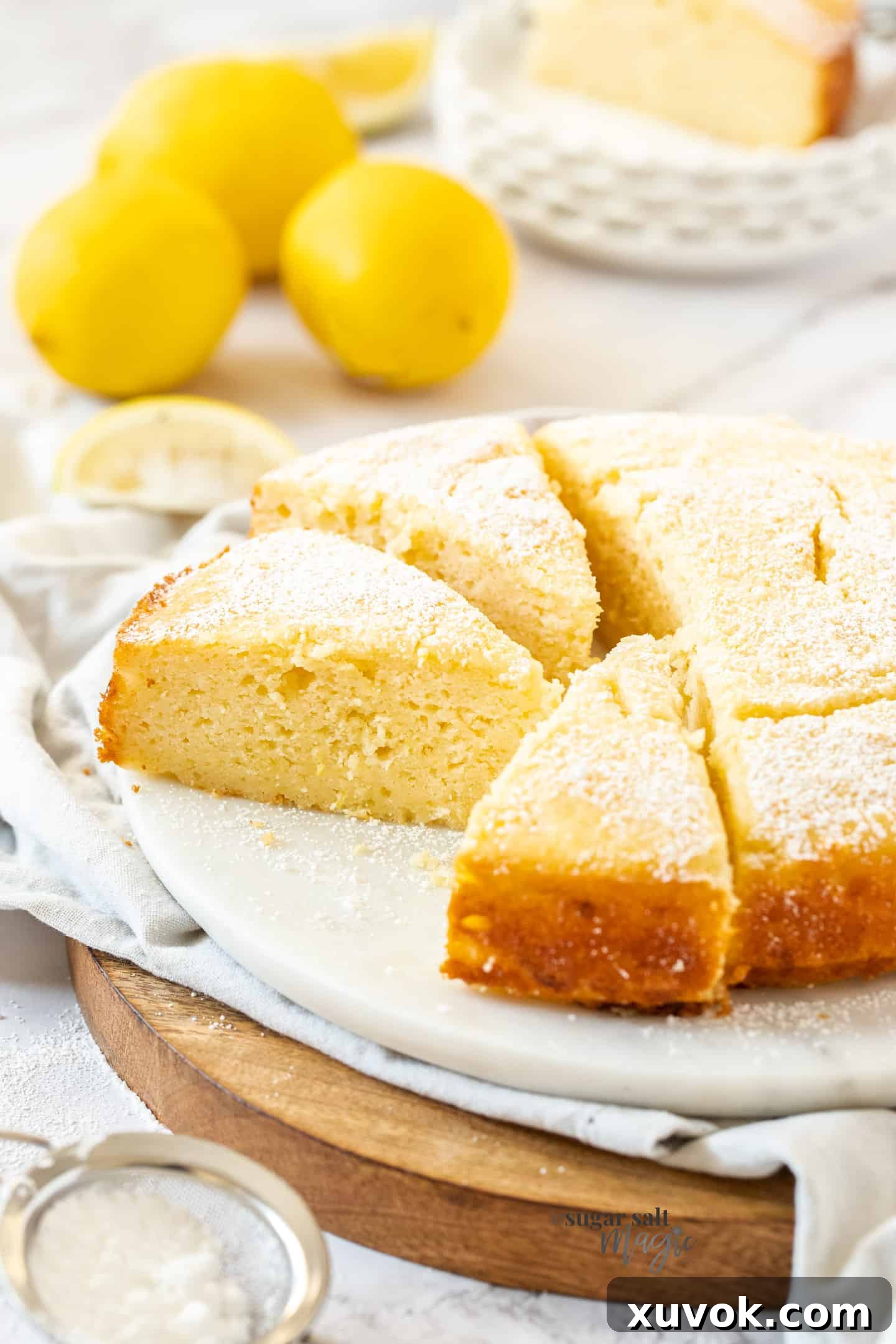 A lemon ricotta cake cut into generous slices on a pristine white platter. Fresh lemons and vibrant green leaves are artfully arranged in the background, highlighting the cake's zesty flavor profile. The cake has a beautiful golden crust and a soft, inviting interior.