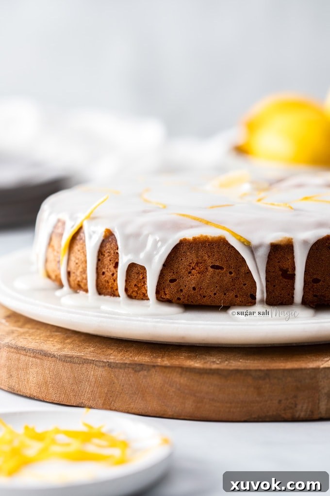 A beautifully decorated lemon cake, generously drizzled with white icing, rests on a pristine marble platter beside a rustic wooden board, with fresh lemons artfully arranged in the background.