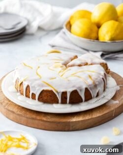 A lemon cake drizzled with white icing on a marble platter and wooden board. Lemons in the background.