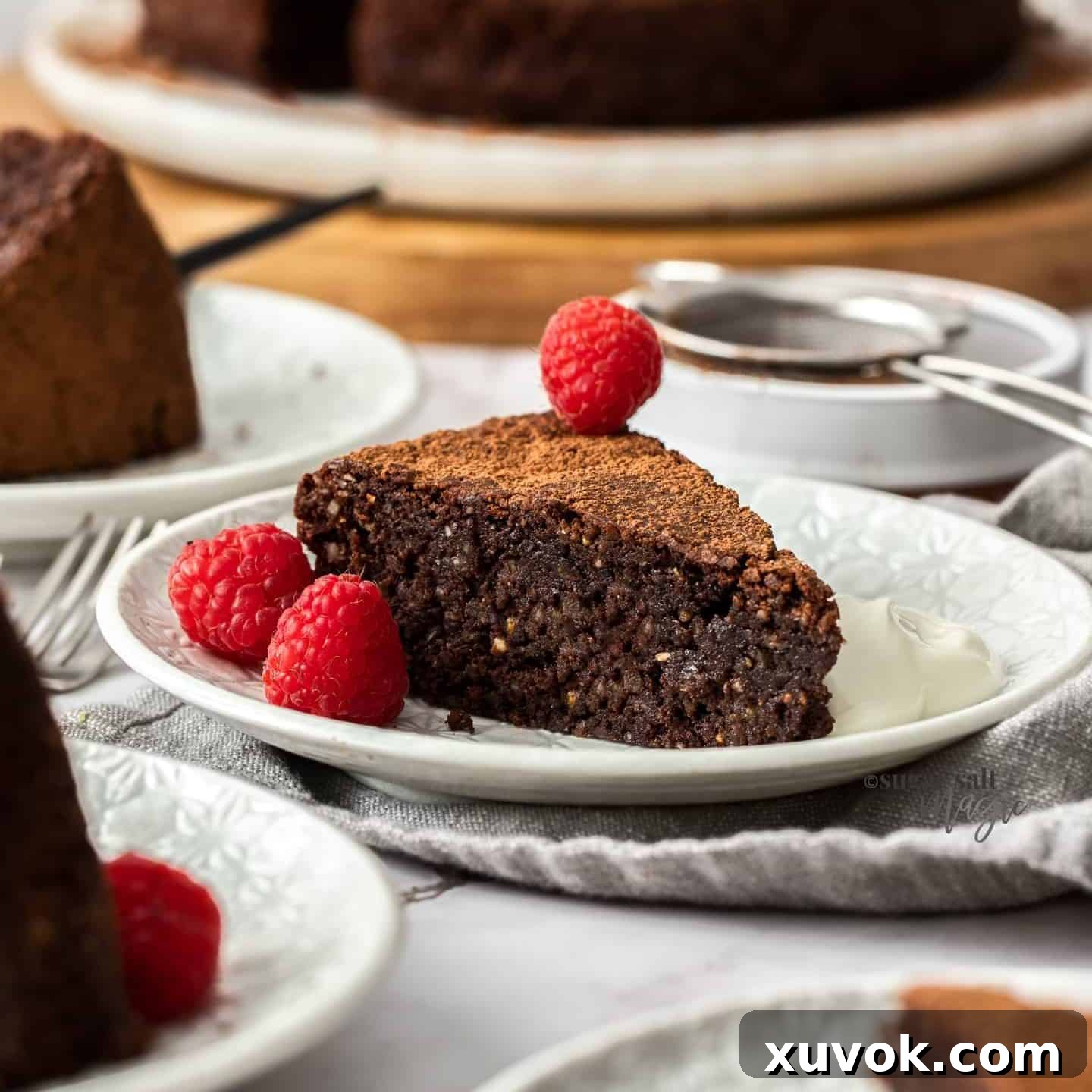 A slice of chocolate cake on a white cake plate with cream and raspberries.