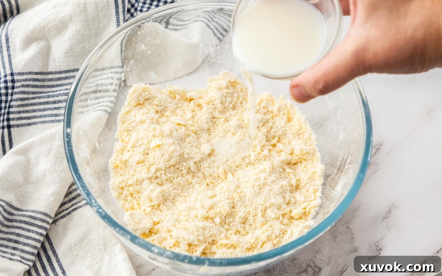 A slow stream of cold milk being carefully poured into the prepared pastry ingredients, starting the process of forming the biscuit dough.