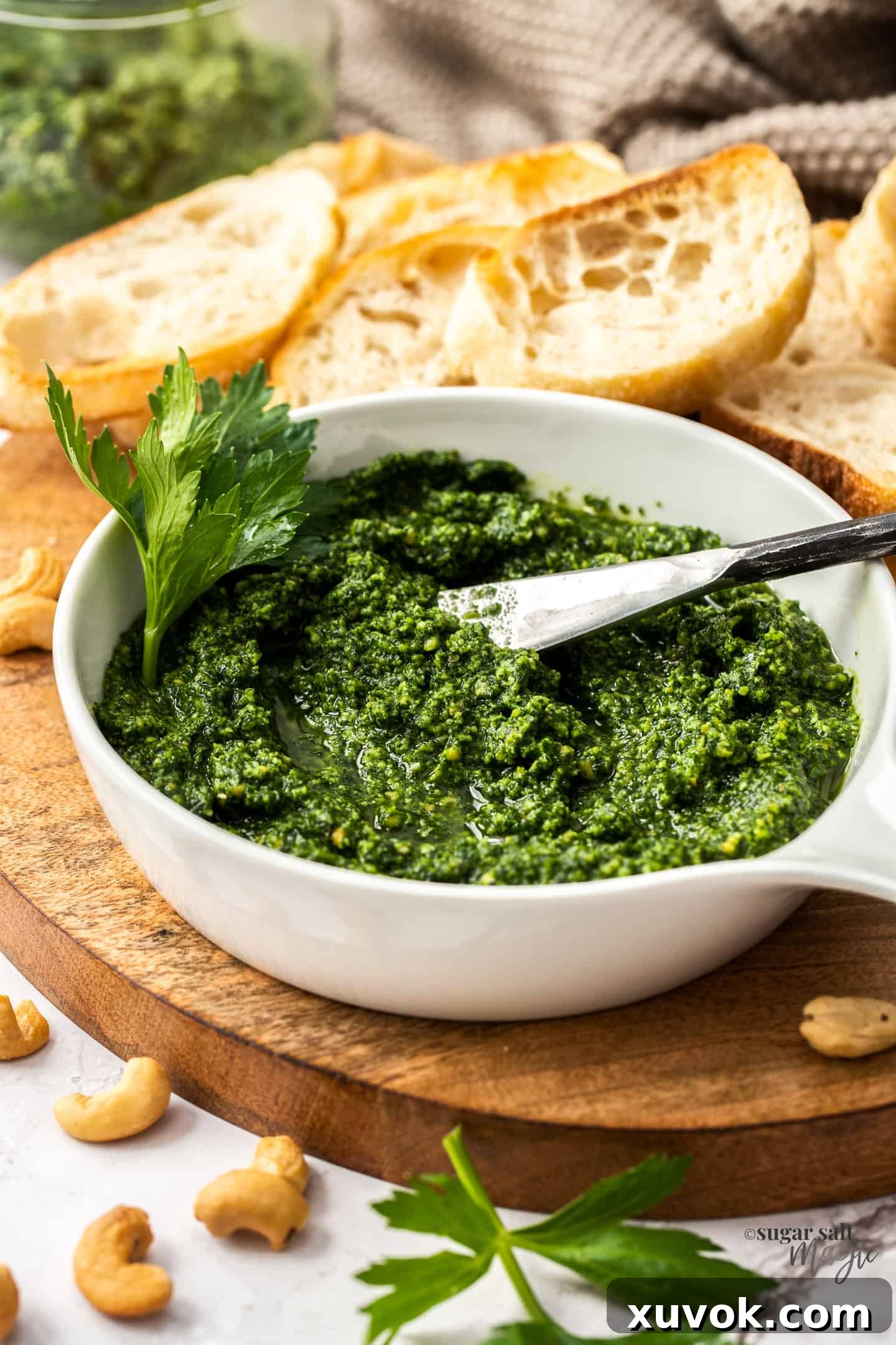 A white bowl filled with vibrant green celery leaf pesto, garnished with a single celery leaf, sitting on a rustic wooden board next to a small spoon.