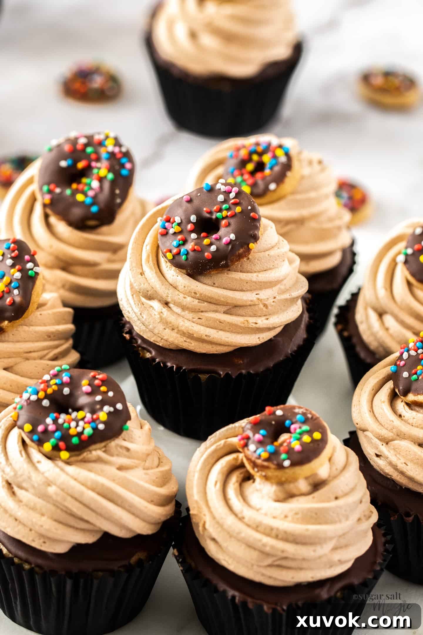 Eight fluffy cinnamon cupcakes with chocolate glaze and whipped chocolate frosting, arranged beautifully on a white marble platter. Each cupcake is adorned with a miniature donut cookie.