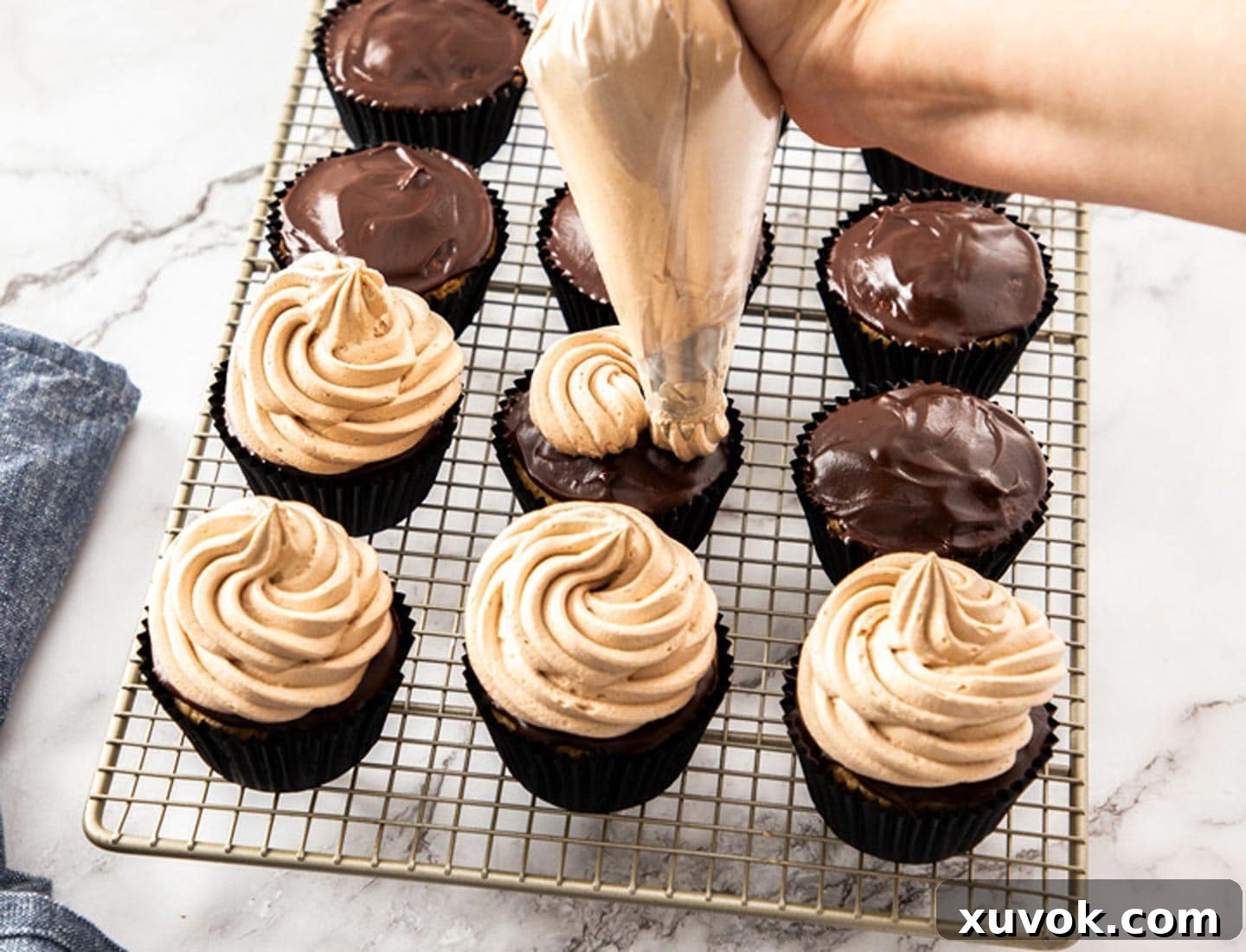 A piping bag elegantly swirling whipped chocolate frosting onto the tops of chocolate-glazed cinnamon cupcakes, creating beautiful mounds.
