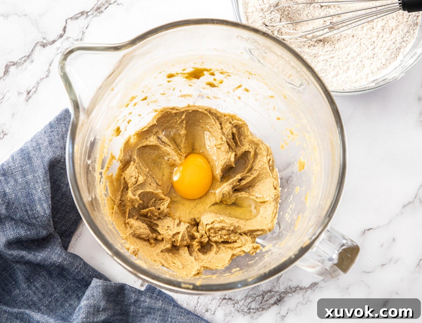 A single egg being cracked and added to the creamy mixture of butter and sugar in a large mixing bowl, ready to be beaten for the cupcake batter.
