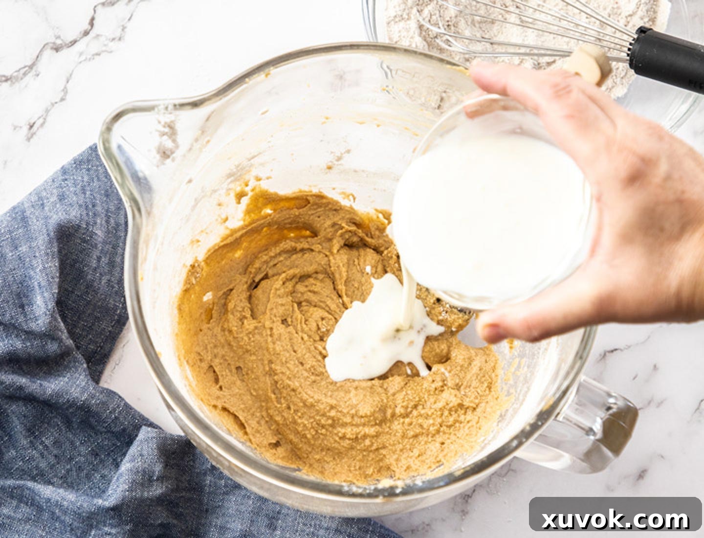 A stream of buttermilk being poured into a fluffy cupcake batter within a large mixing bowl, with a spatula poised to gently combine the ingredients.