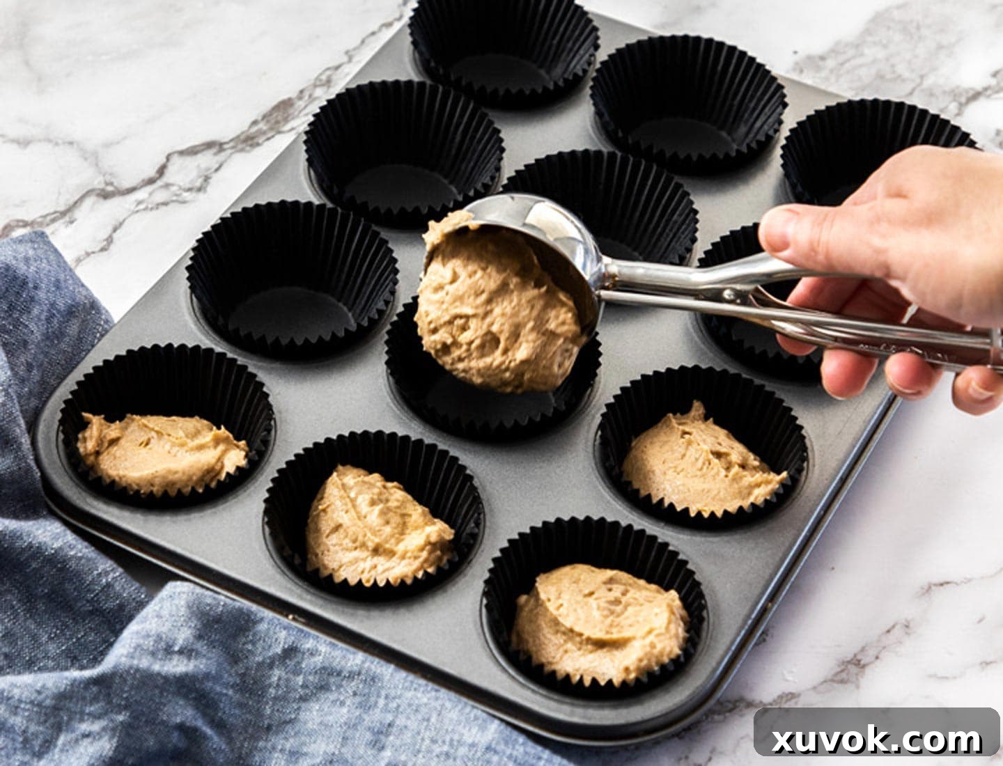 Fluffy cinnamon cupcake batter being scooped from a bowl with an ice cream scoop and carefully tipped into cupcake liners arranged in a baking tray.