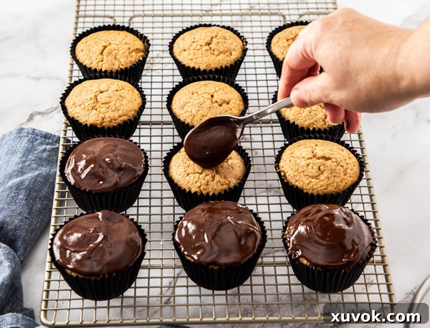 A rich chocolate glaze being carefully spooned onto the golden brown tops of cooled cinnamon cupcakes, creating a smooth and shiny layer.
