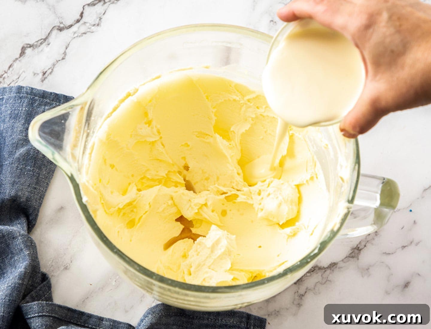 A generous pour of fresh whipping cream being added to the light and fluffy whipped butter and sugar mixture in a stand mixer bowl.