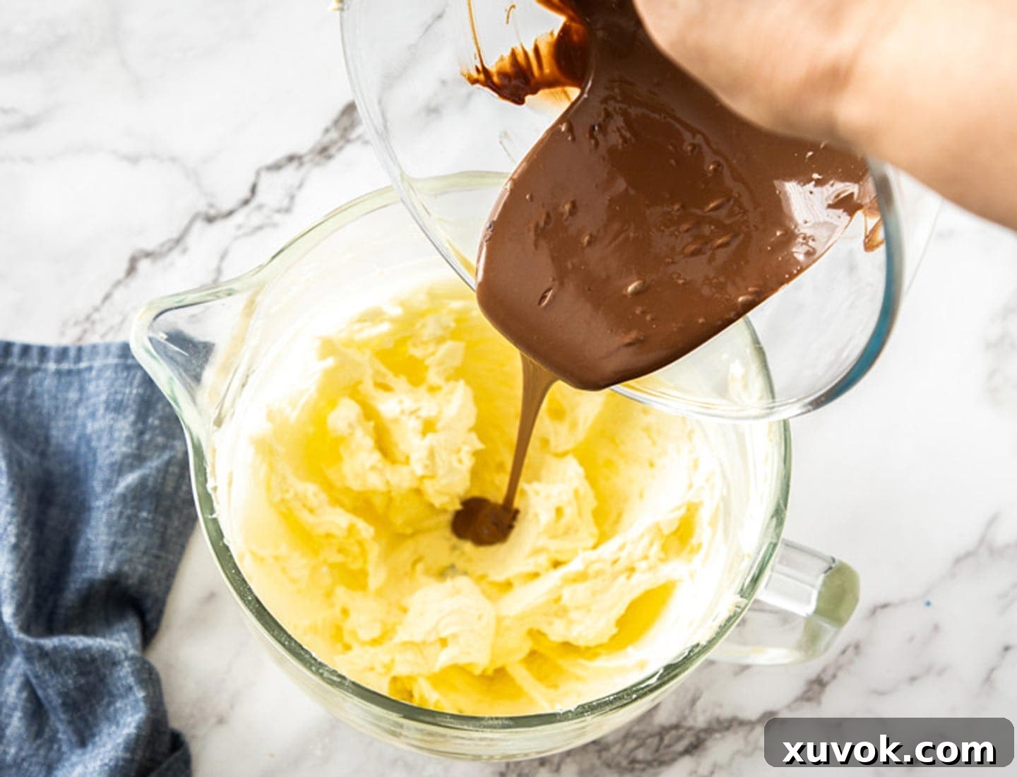 Stream of melted milk chocolate being poured into a bowl of already whipped butter and sugar, preparing for the final stage of frosting creation.
