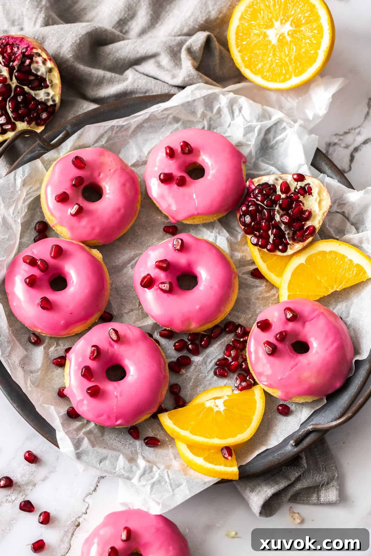 Six vibrant orange donuts adorned with bright pink pomegranate icing, artfully arranged amidst fresh orange slices and ruby-red pomegranate seeds on a light background. The image exudes freshness and deliciousness, highlighting the donut's appealing colors and textures.