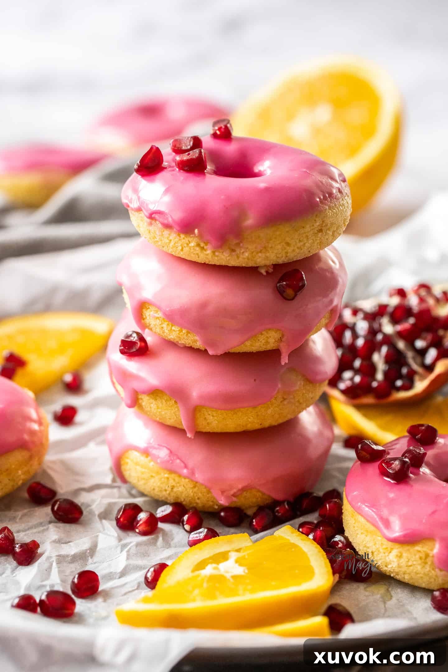 A beautifully composed stack of four orange donuts, generously coated with glistening pink pomegranate icing, surrounded by scattered ruby-red pomegranate seeds on a rustic surface. The donuts appear fresh and inviting, ready to be enjoyed.