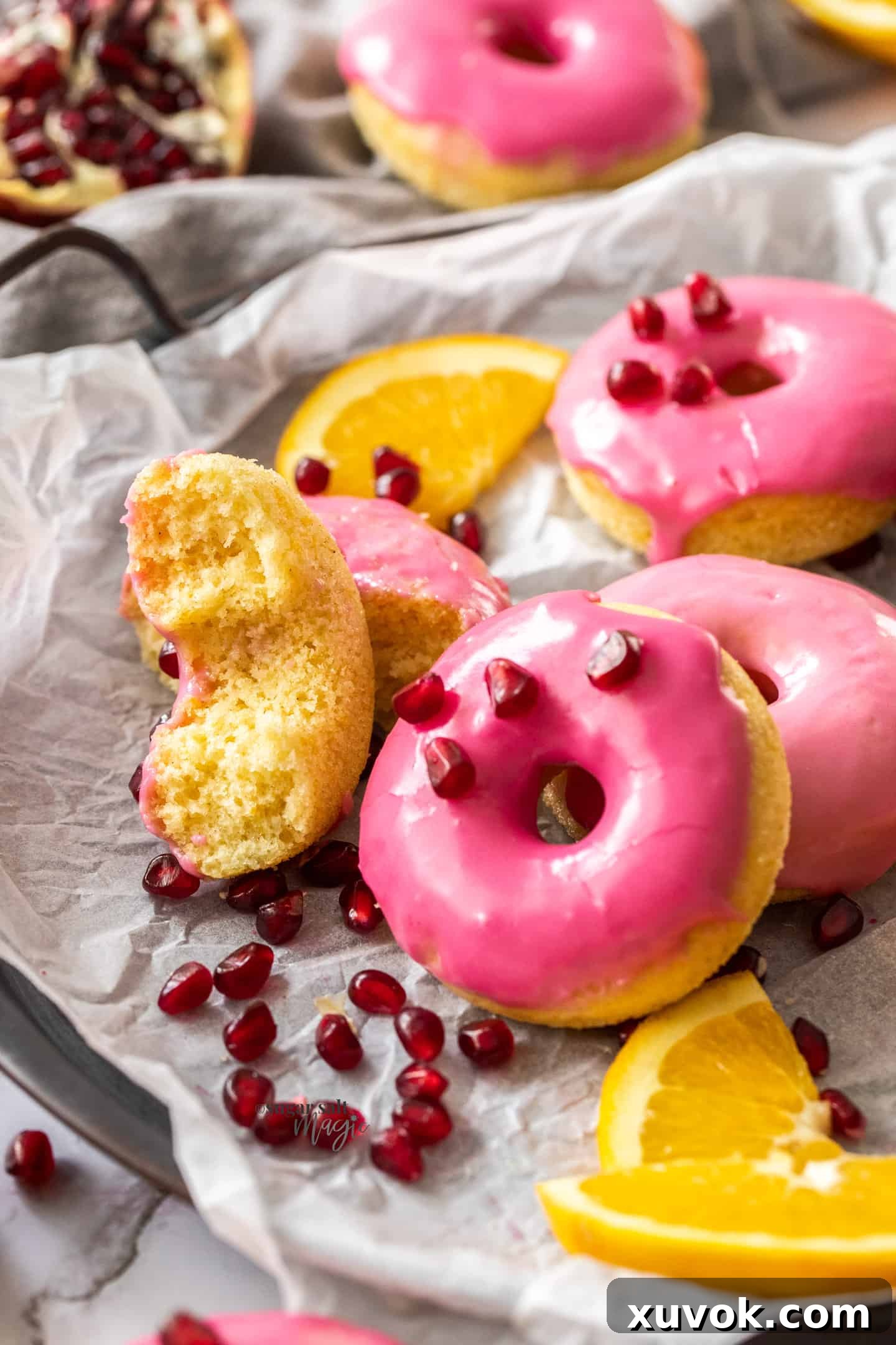 A delectable baked orange donut, adorned with pink pomegranate icing, gently broken open to reveal its moist, tender interior. It rests on a sheet of baking paper, surrounded by other iced donuts, inviting a taste.