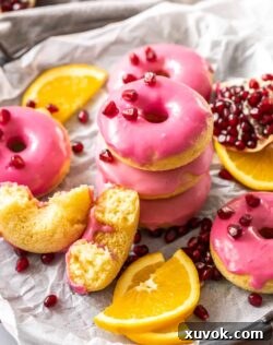 A stack of beautifully iced orange donuts, with a vibrant pink pomegranate glaze, surrounded by fresh orange slices and ruby pomegranate seeds, presenting a delightful and inviting treat.