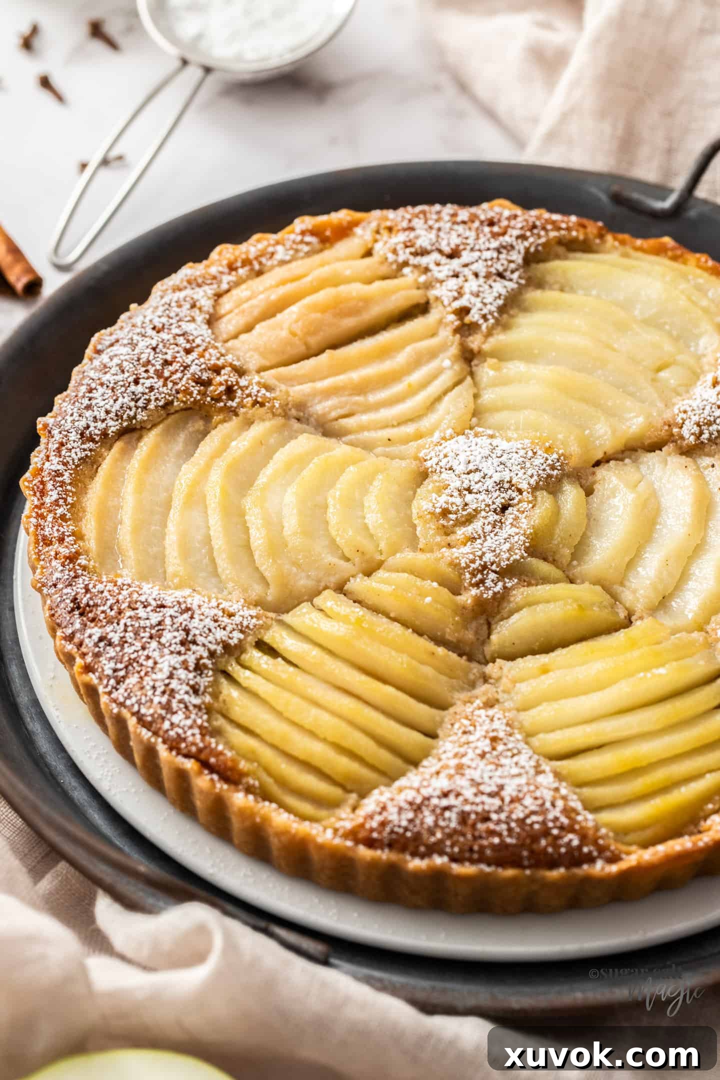 A tart filled with slices of pears on a white cake plate. A small sieve of icing sugar in the background.