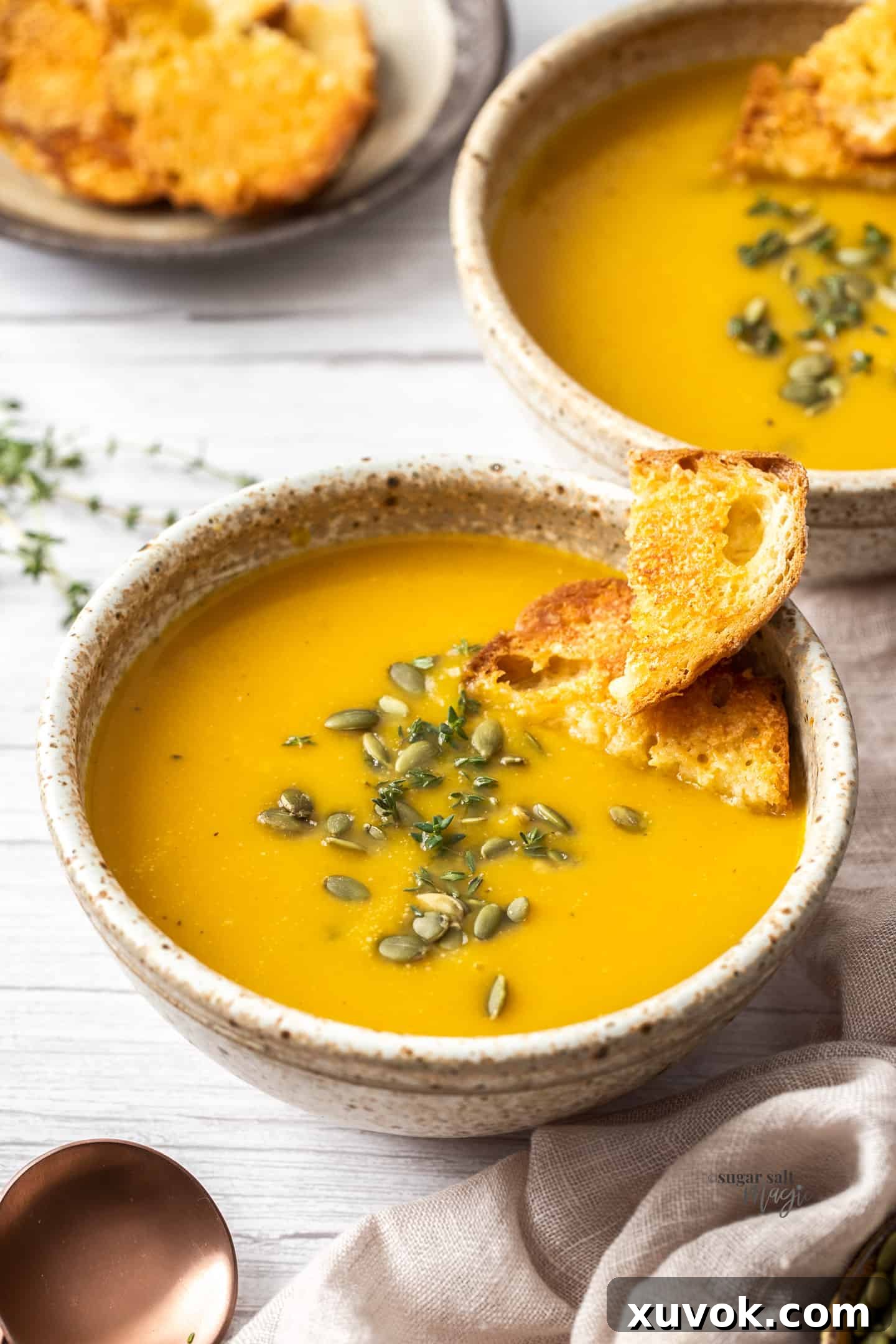 A bowl filled with vibrant orange butternut squash soup, garnished with fresh herbs and pepitas, served alongside a slice of crusty bread for dipping. Another similar bowl is softly blurred in the background.