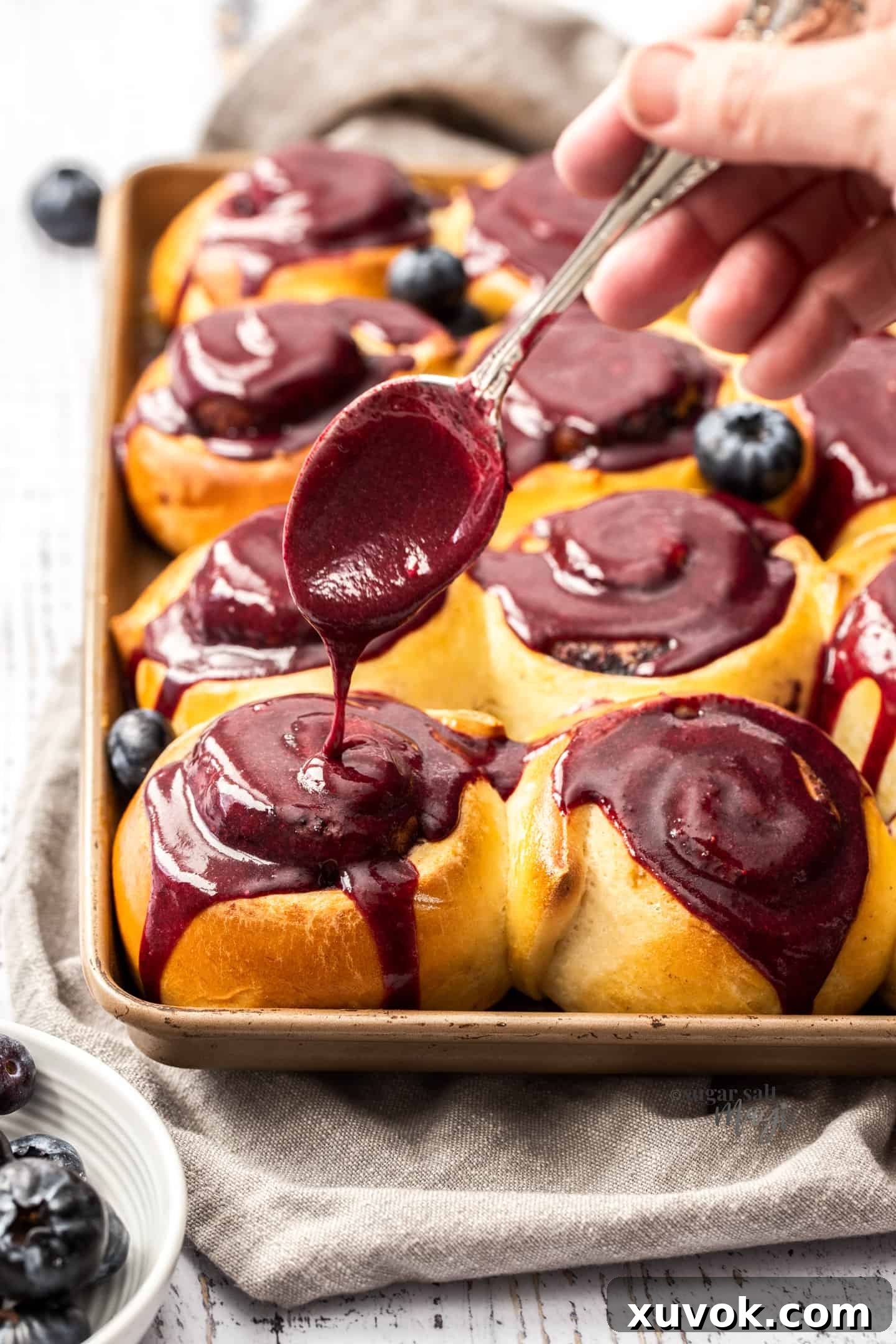 Blueberry glaze being drizzled generously over the top of warm cinnamon rolls in a baking tin with a spoon, highlighting their rich blue color and soft texture.