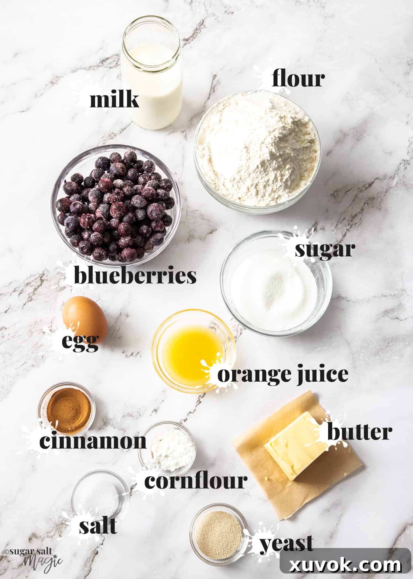 All the fresh ingredients for homemade blueberry cinnamon rolls, neatly arranged on a pristine marble surface, including flour, butter, milk, sugar, eggs, yeast, fresh blueberries, orange juice, and cinnamon.