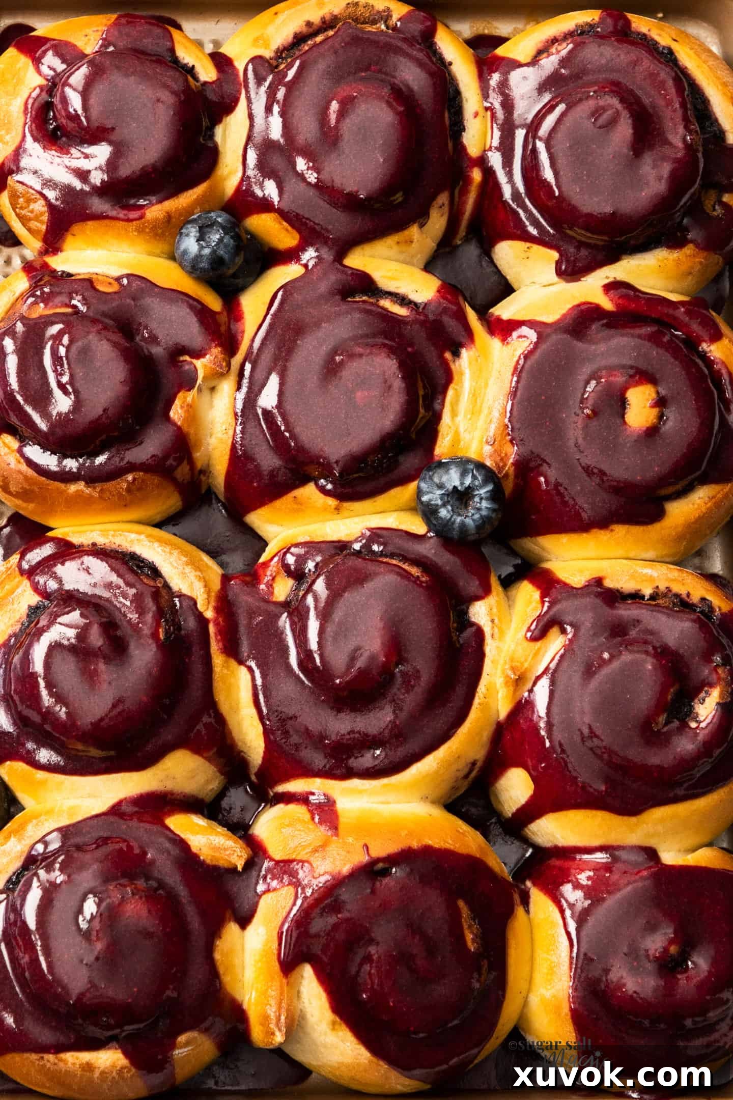 A top-down, close-up view of 12 freshly baked blueberry cinnamon rolls, glistening with a generous layer of vibrant blueberry glaze, still arranged in their baking tray.