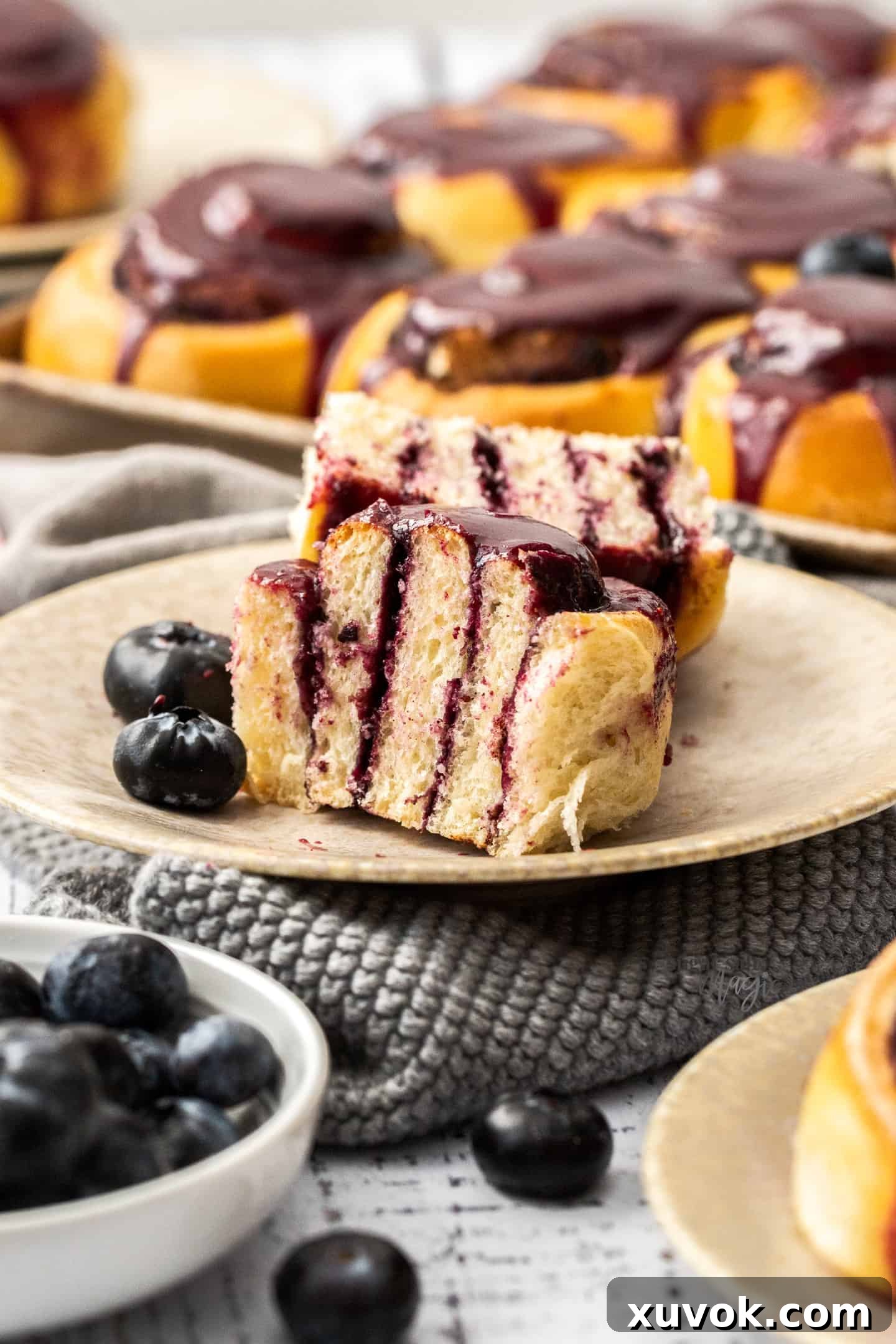 A single, perfectly baked blueberry cinnamon roll, adorned with vibrant blueberry glaze, presented on a small cream-colored plate and cut in half to reveal its soft, swirled interior and jammy filling.