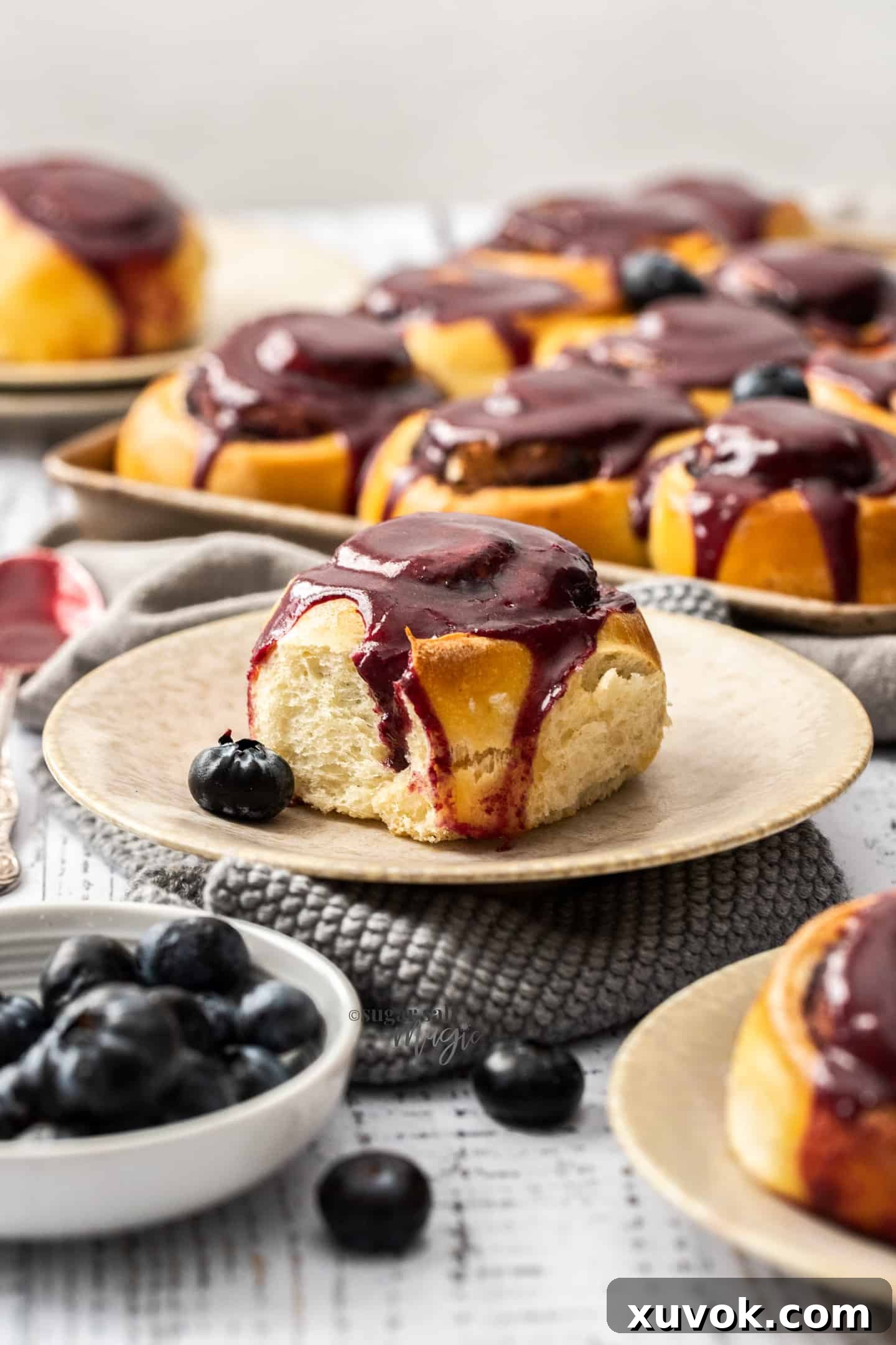 A single, delectable blueberry cinnamon roll, generously topped with a bright purple blueberry glaze, artfully placed on a pristine cream-colored plate, ready to be enjoyed.