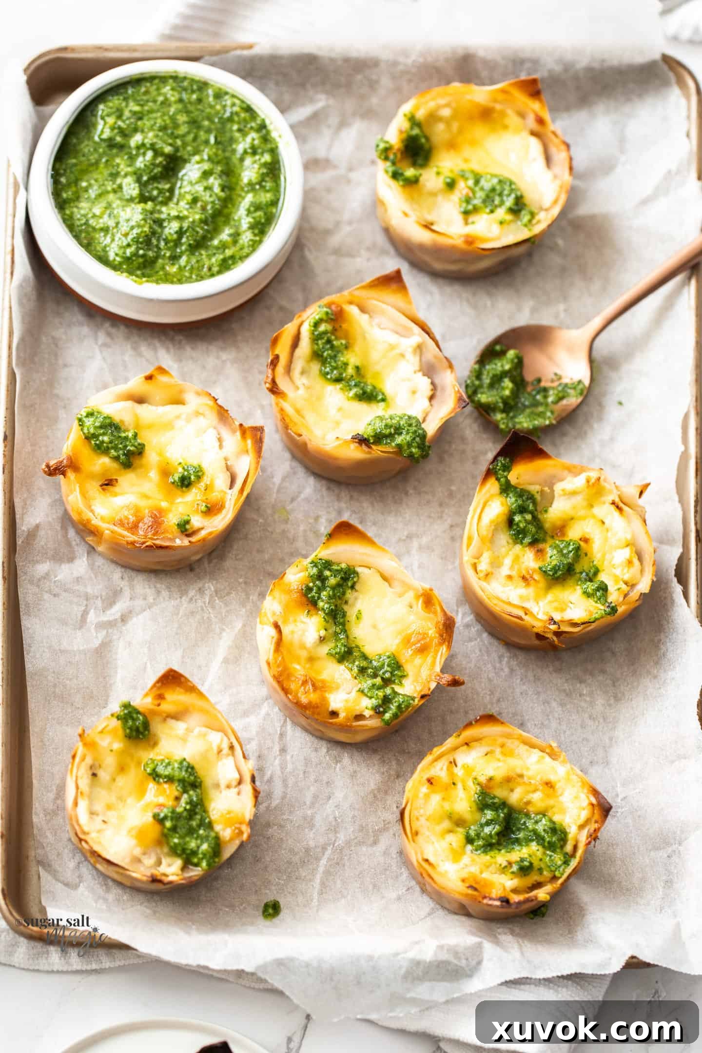 Seven golden-brown lasagna cupcakes arranged neatly on a baking paper-lined tray, accompanied by a small bowl of vibrant green pesto, highlighting their appealing presentation and delicious potential as party food or an Italian appetizer.