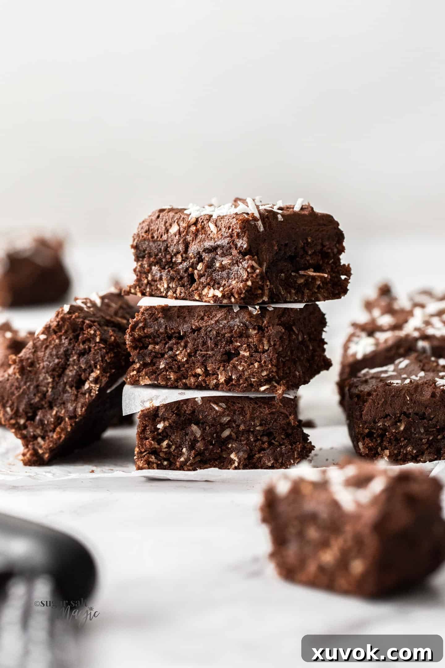 A stack of 3 pieces of chocolate coconut slice topped with shredded coconut, showcasing its fudgy texture and delicious chocolate frosting.