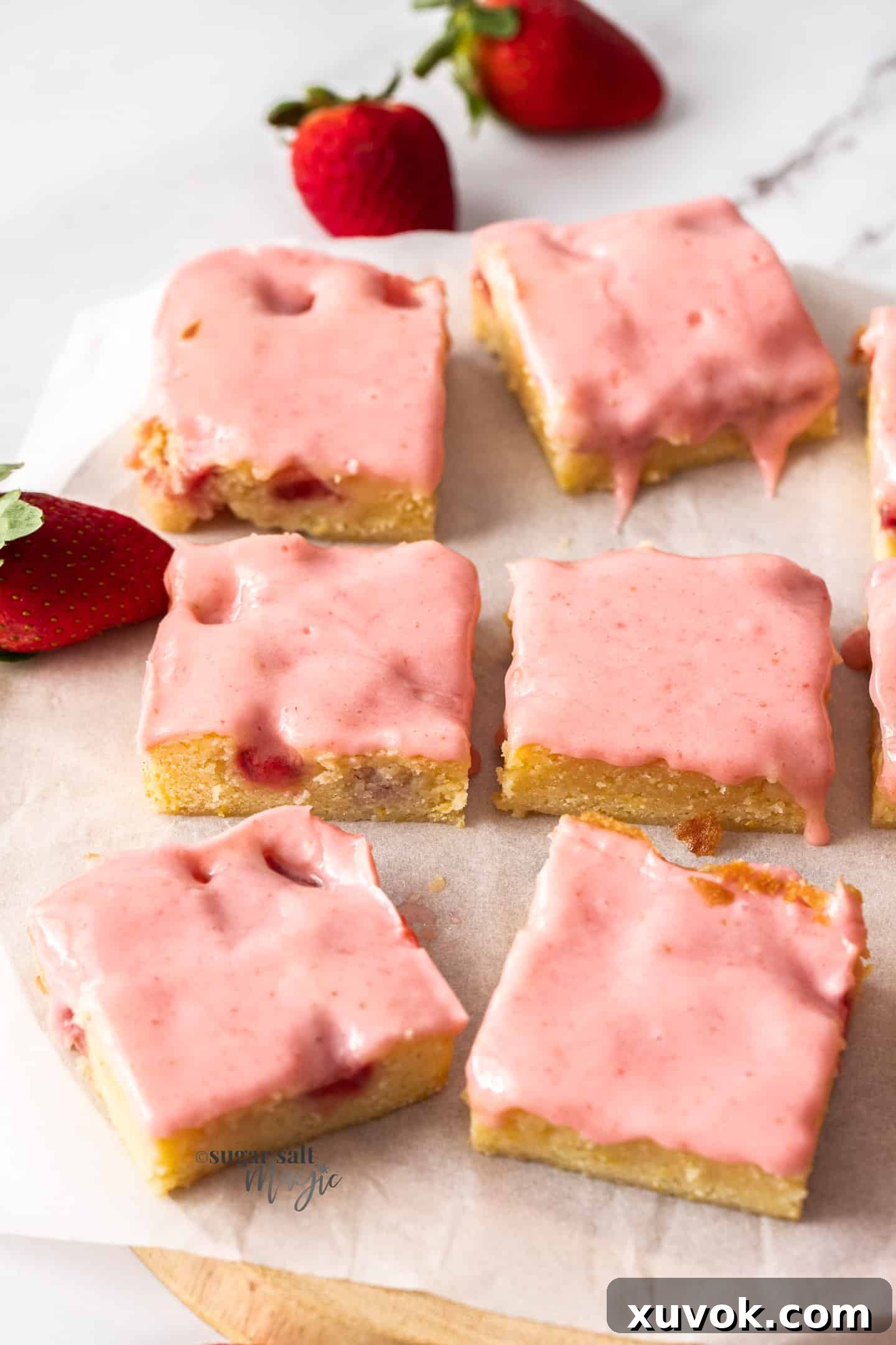 Six square blondies with pink strawberry icing on a sheet of white baking paper, arranged for a tempting display.