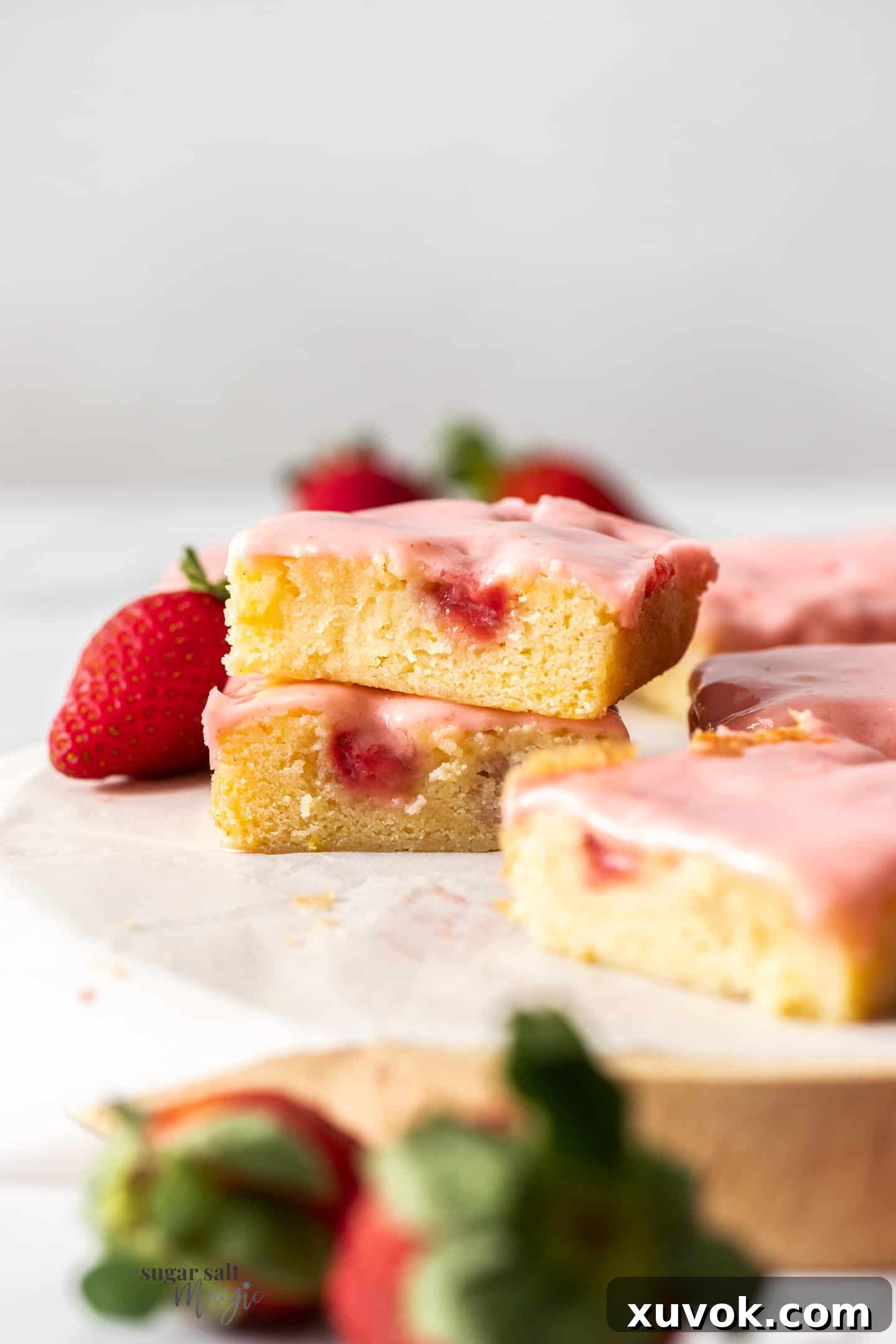 A close-up shot of two strawberry blondies stacked, with more blondies and a fresh strawberry in the background, highlighting the moist texture and pink icing.