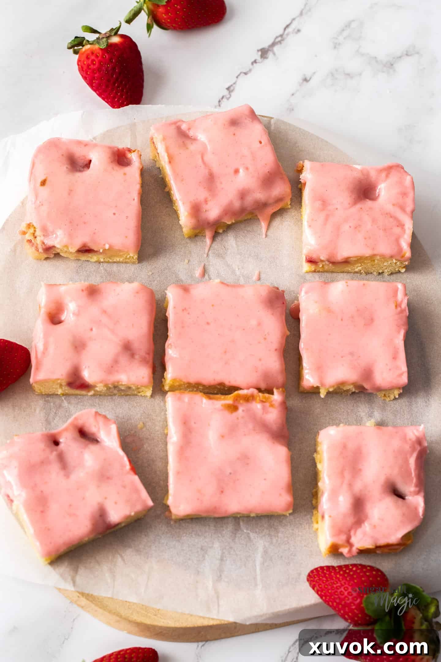 Nine perfectly cut blondies with luscious pink icing displayed on a sheet of baking paper, set on a rustic wooden board, ready to be enjoyed.