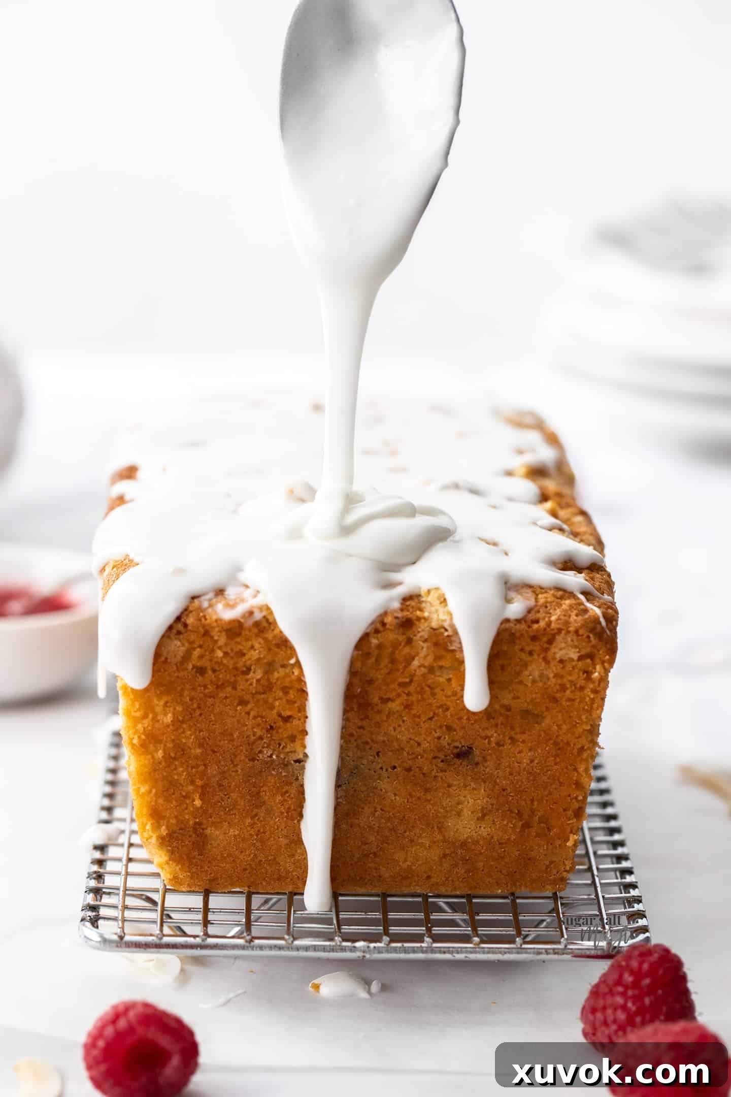 A spoon gently drizzling smooth white coconut icing directly down onto a freshly baked coconut loaf cake, creating elegant trails.