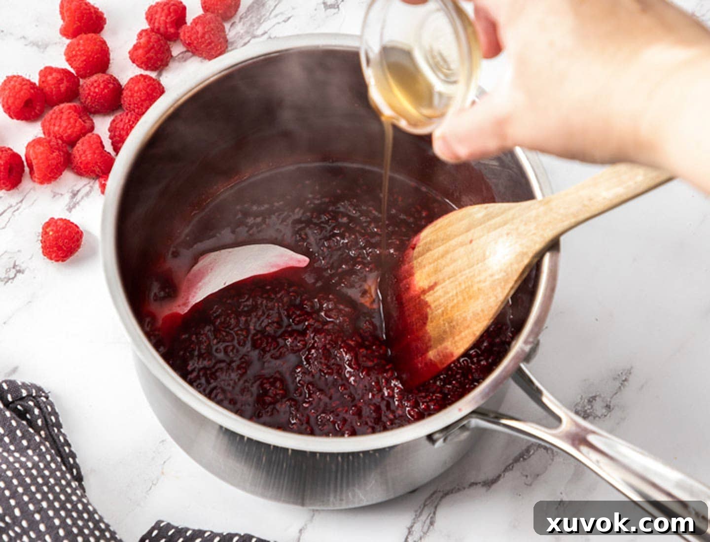 A hand pouring vanilla extract from a small bowl into a saucepan of simmering raspberry compote, creating aromatic steam.
