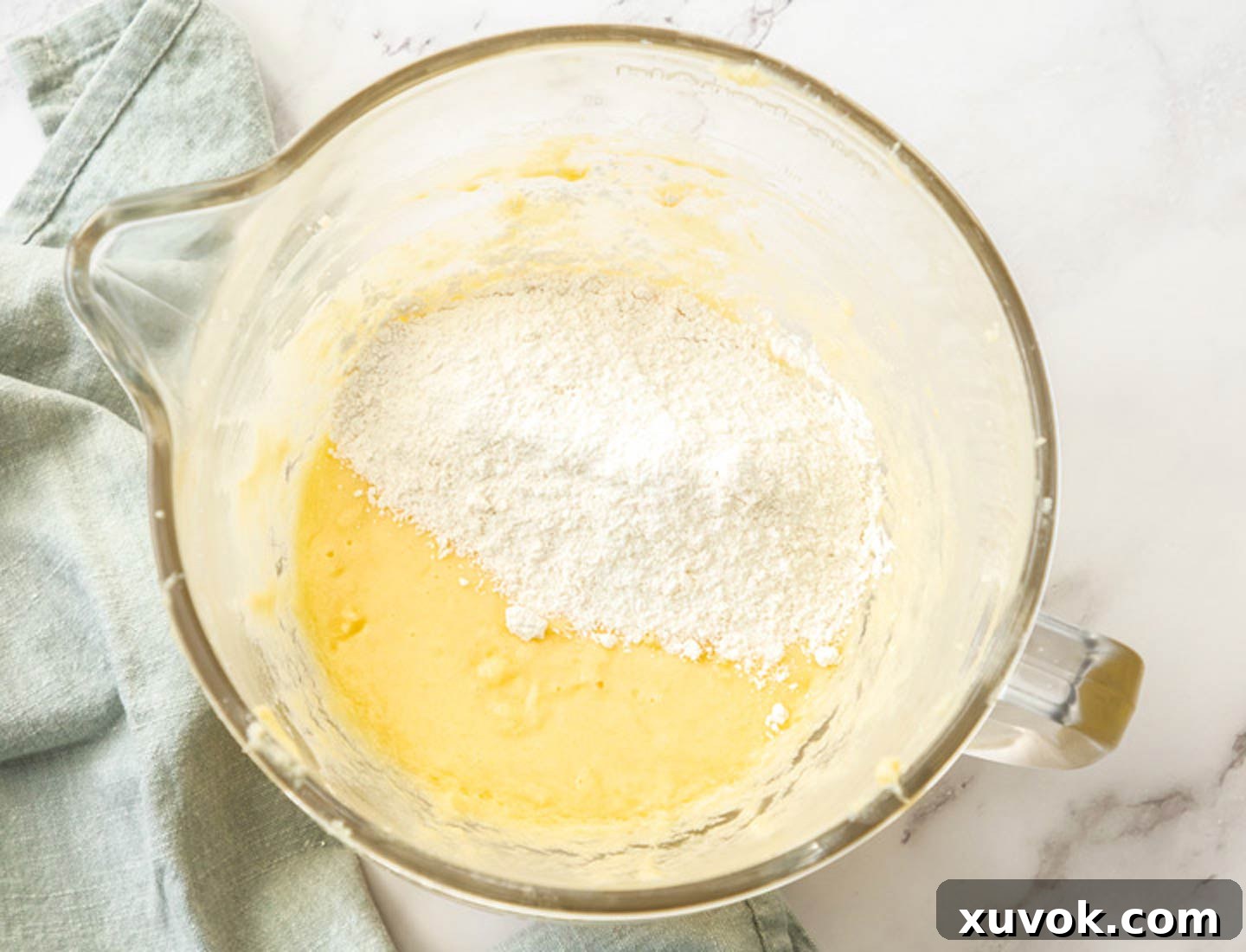 The dry ingredients, a blend of flour, desiccated coconut, baking powder, and salt, are gently folded into the creamy wet ingredients in a large mixing bowl.
