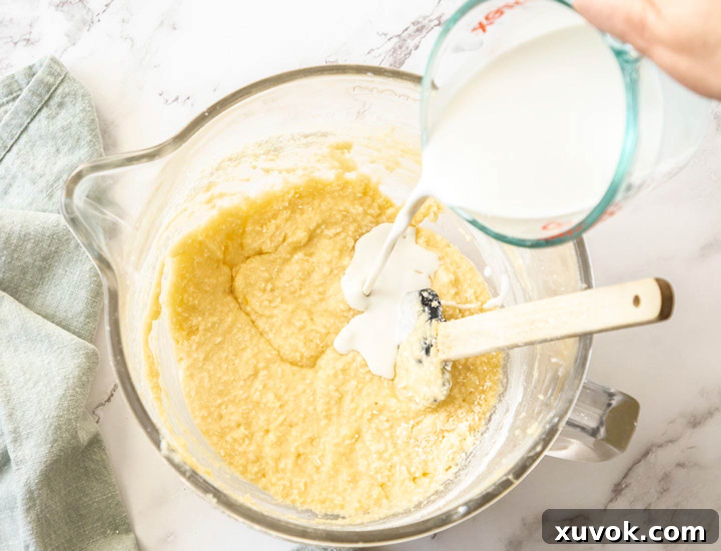 A stream of rich coconut milk being poured into the cake batter in a clear glass mixing bowl, ready to be gently folded in.