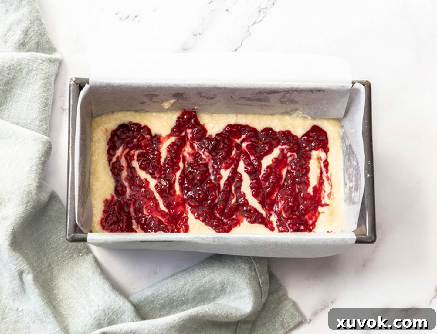 Vibrant raspberry compote being delicately swirled over a layer of cake batter in a baking pan, creating beautiful patterns.