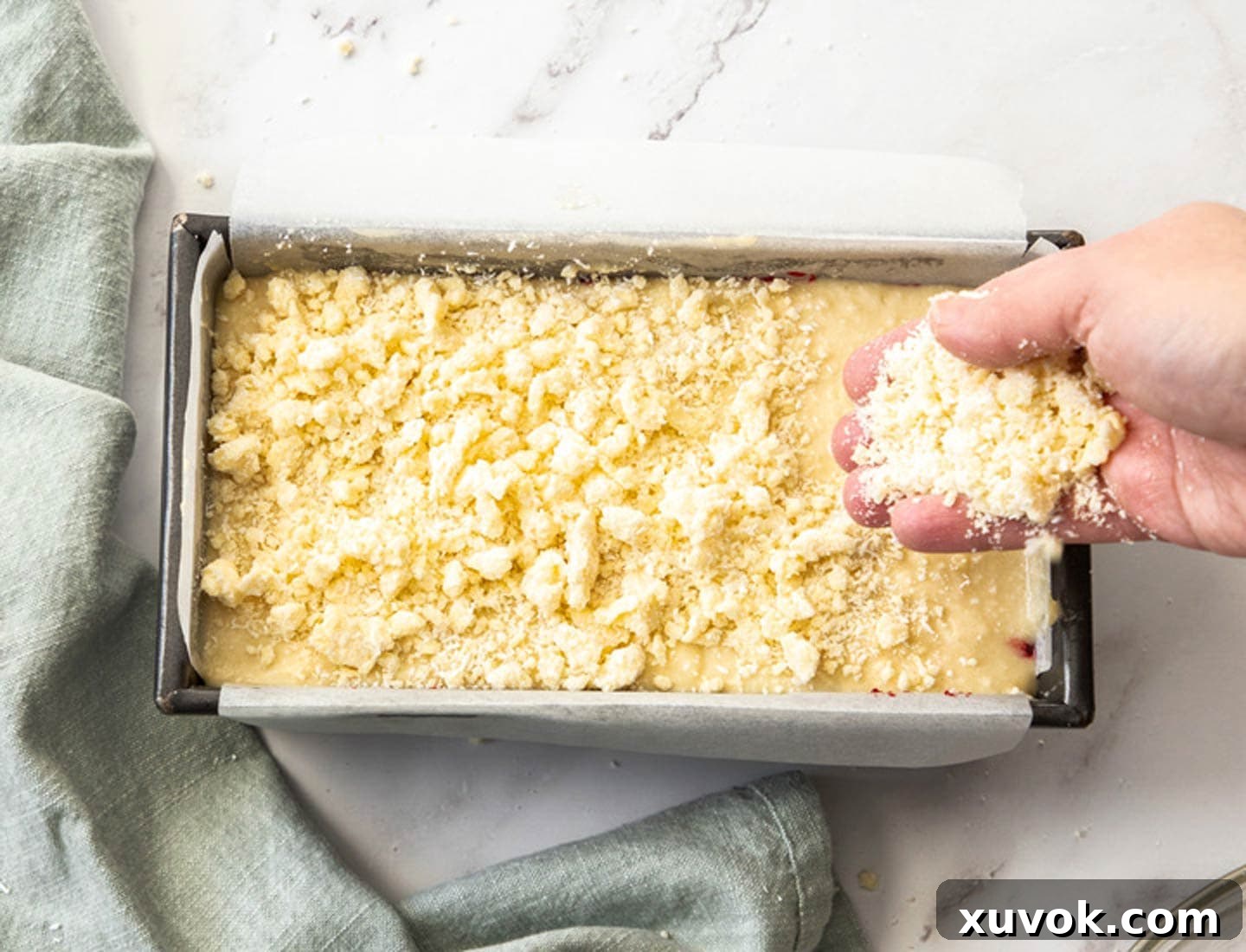 A hand scattering golden-brown coconut crumble evenly over the top of the cake batter in a baking pan, preparing it for the oven.